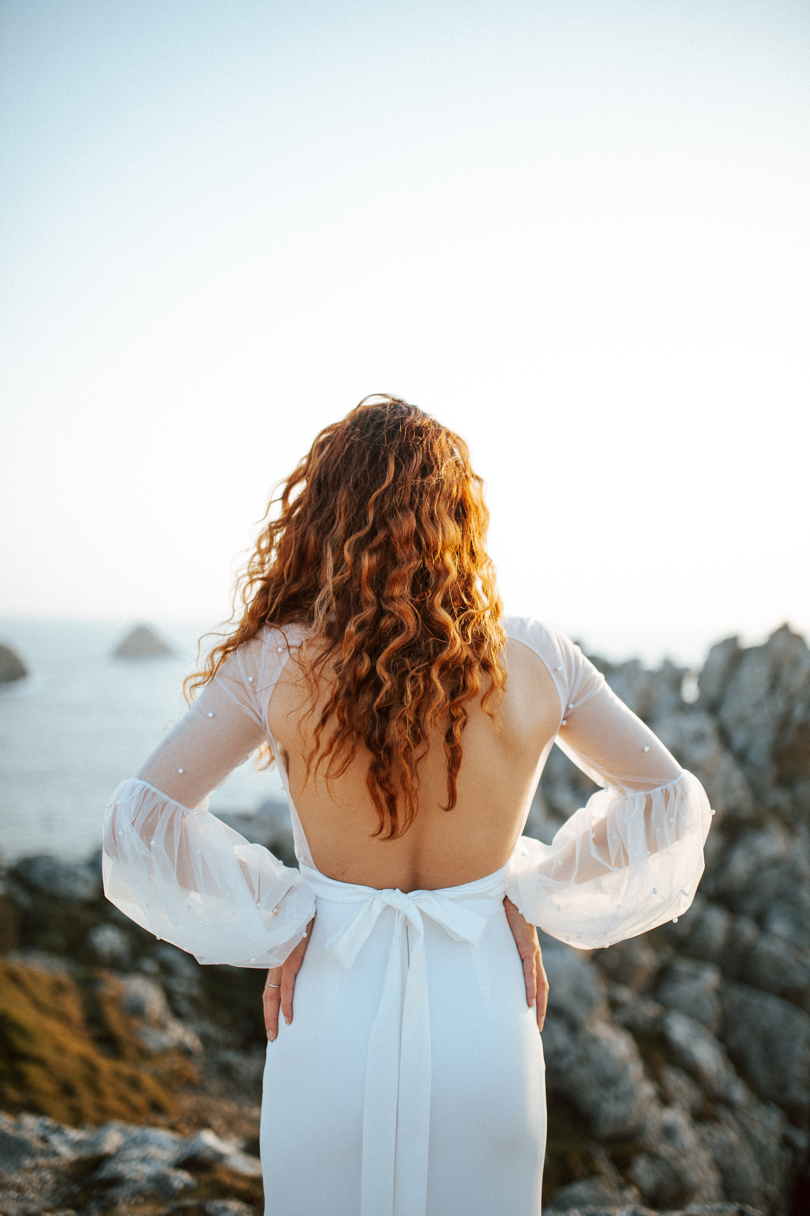 Femme rousse portant une robe de mariée brodée de perles en bord de mer.