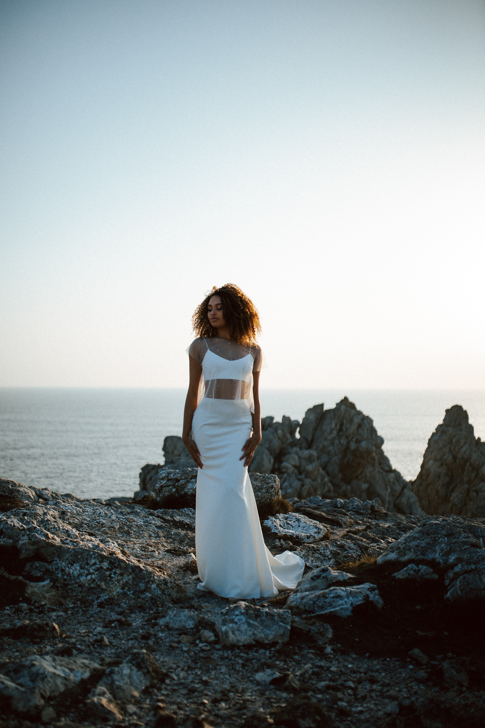 Femme métissée portant une robe de mariée brodée de perles en bord de mer.