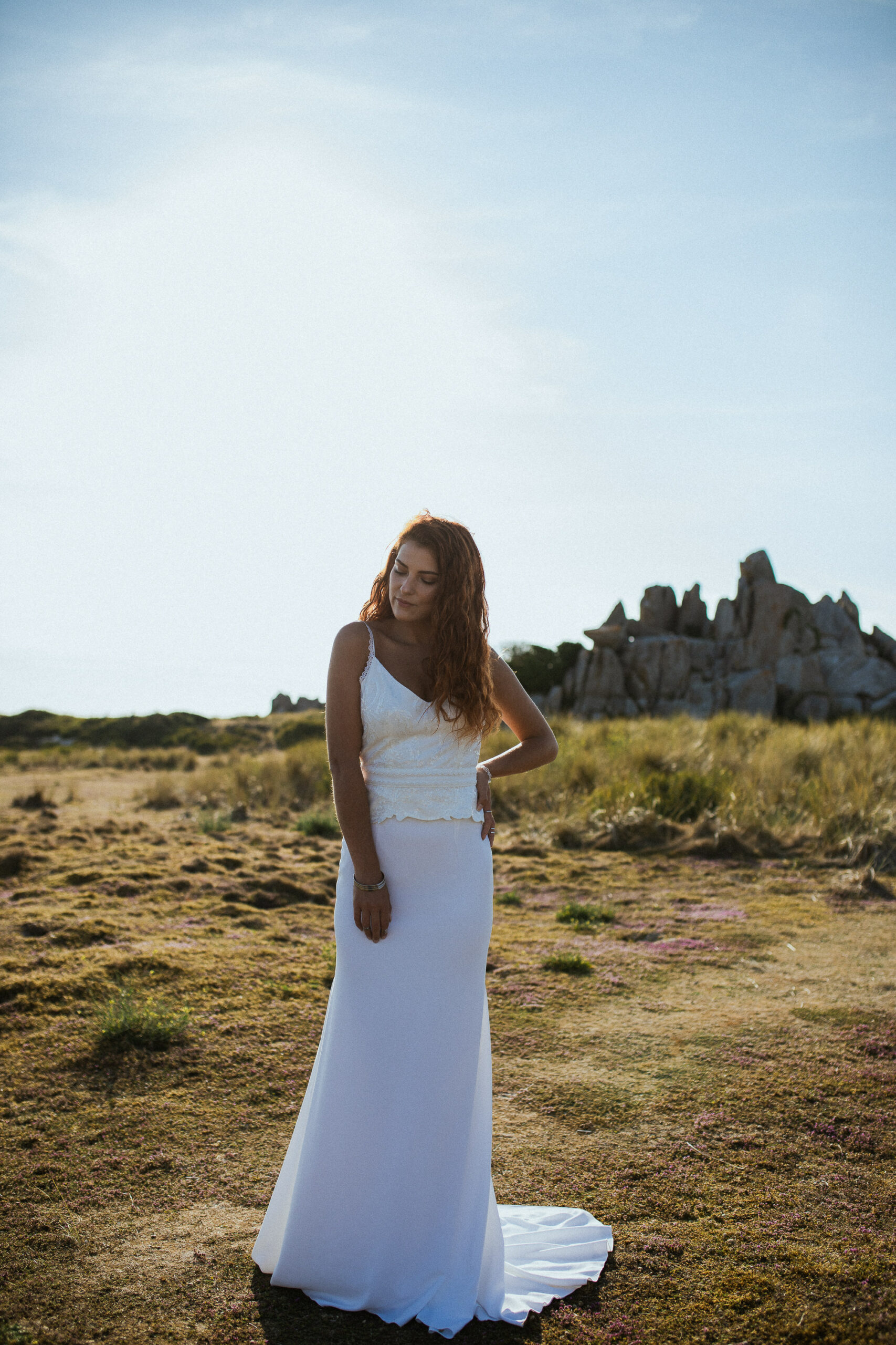 Femme rousse portant une robe de mariée en dentelle dans les dunes végétales en bord de mer.