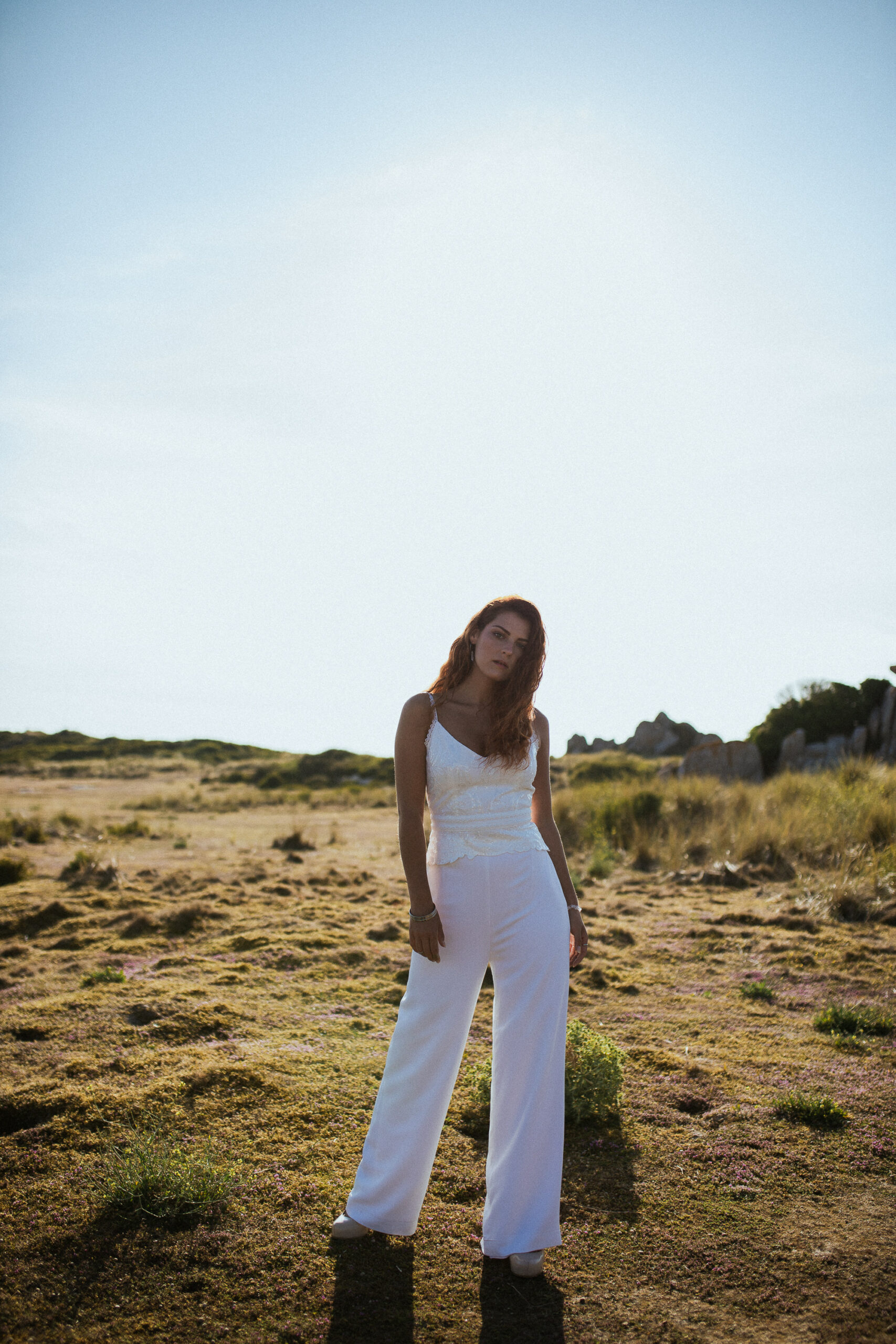 Femme rousse portant une robe de mariée en dentelle dans les dunes végétales en bord de mer.