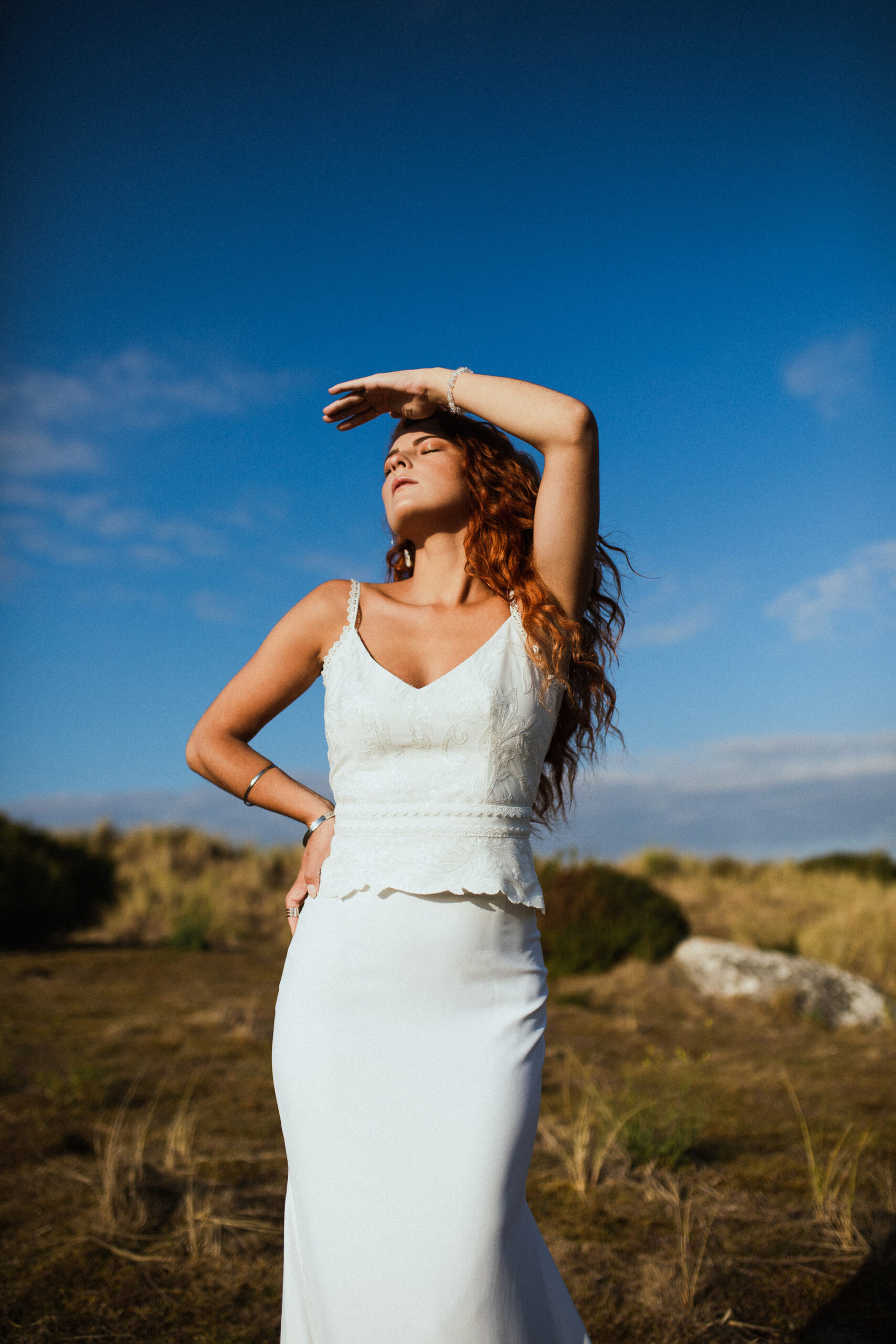 Femme rousse portant une robe de mariée en dentelle dans les dunes végétales en bord de mer.