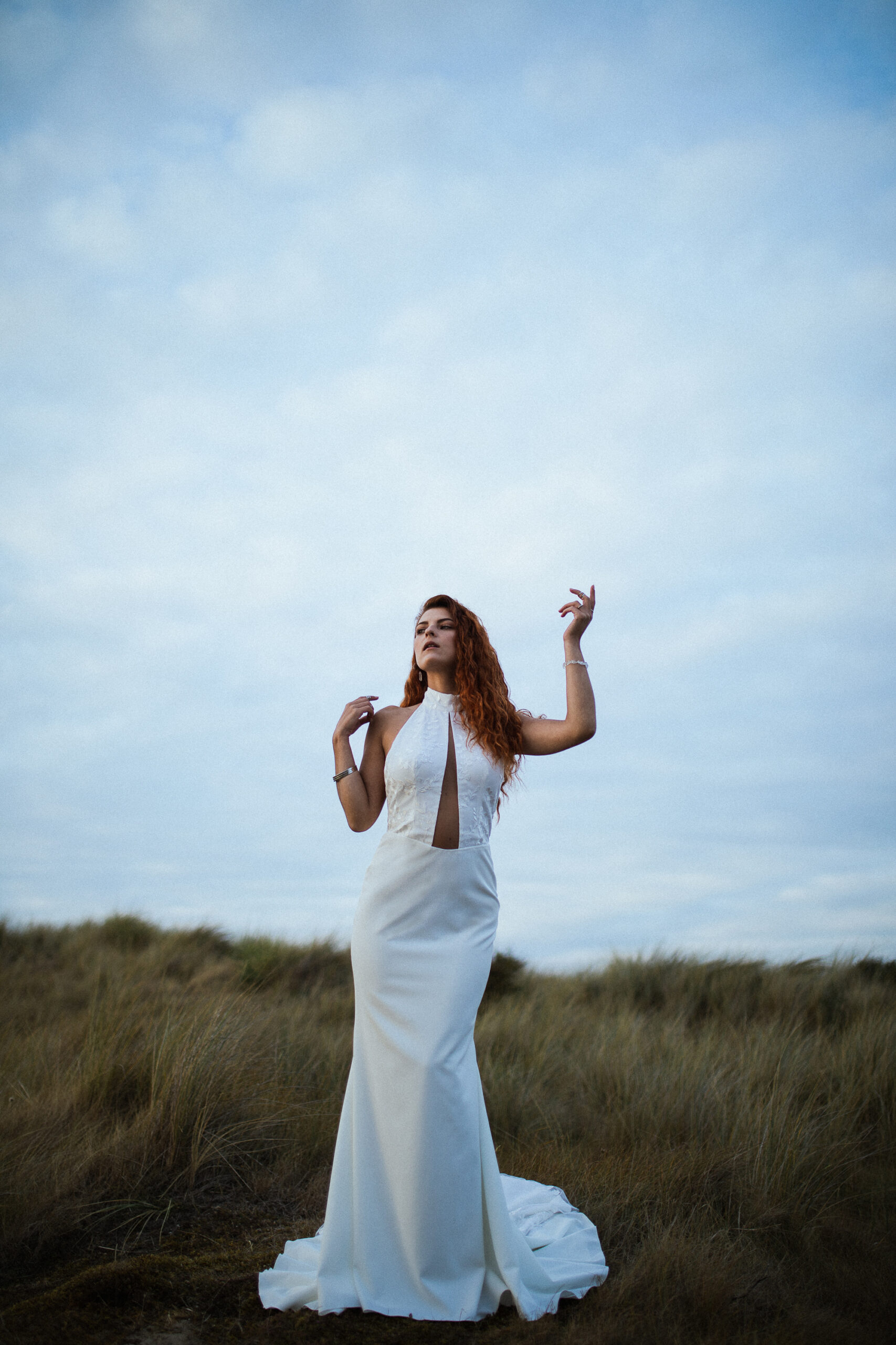 Femme rousse portant une robe de mariée en dentelle dans les dunes végétales en bord de mer.