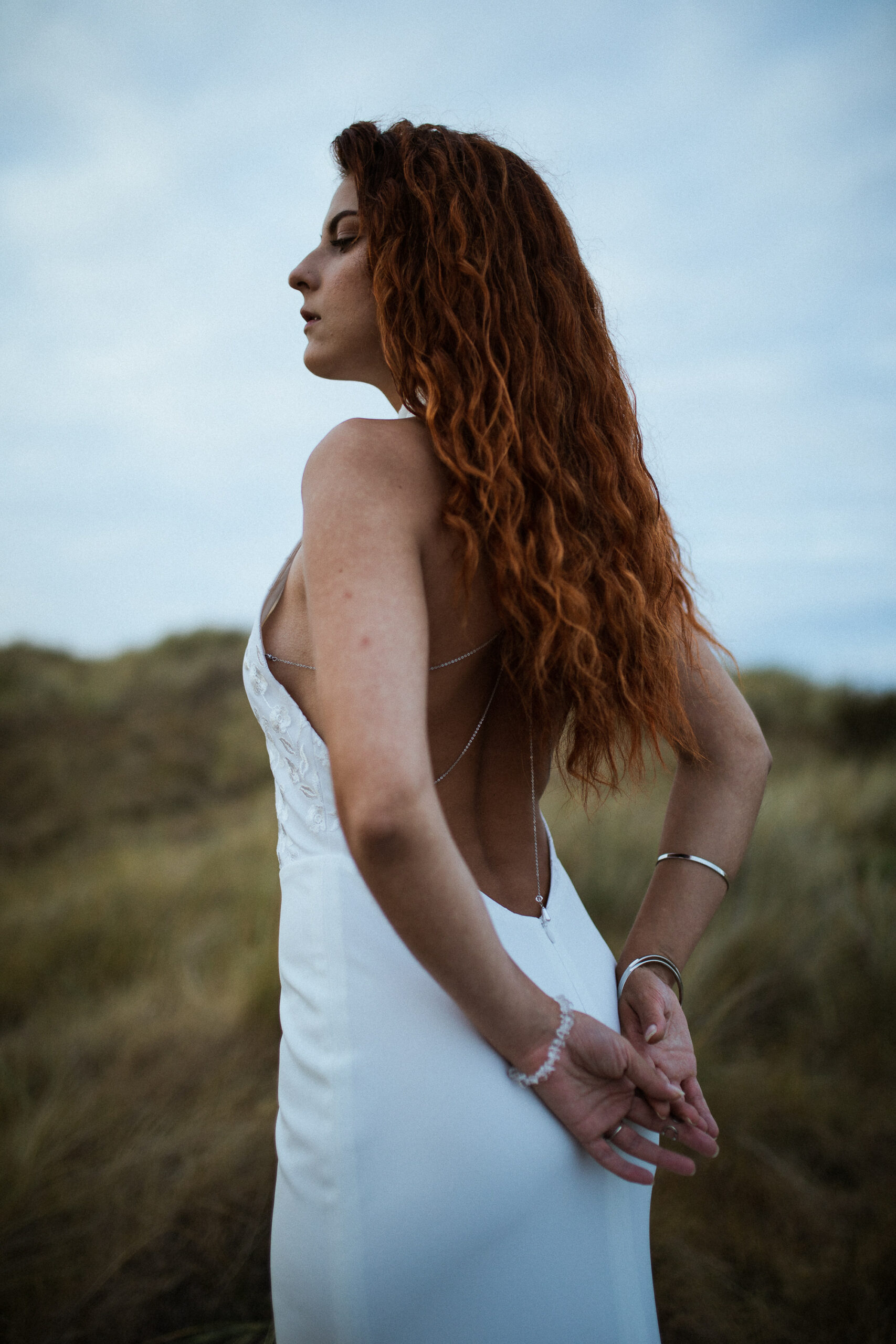 Femme rousse portant une robe de mariée en dentelle dans les dunes végétales en bord de mer.