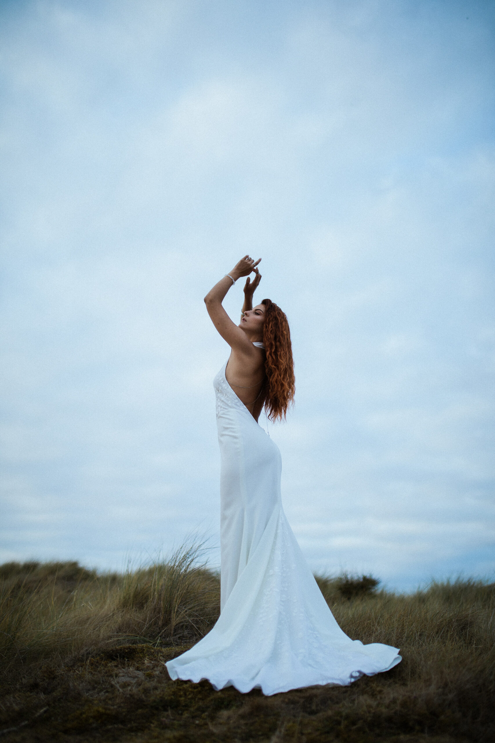 Femme rousse portant une robe de mariée en dentelle dans les dunes végétales en bord de mer.