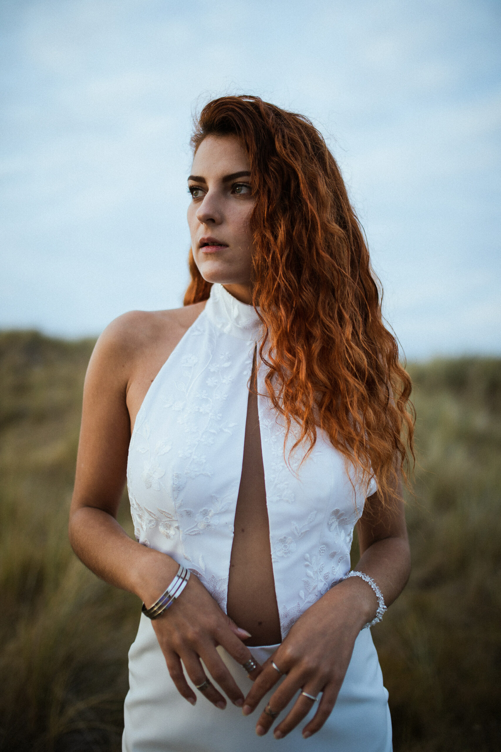 Femme rousse portant une robe de mariée en dentelle dans les dunes végétales en bord de mer.