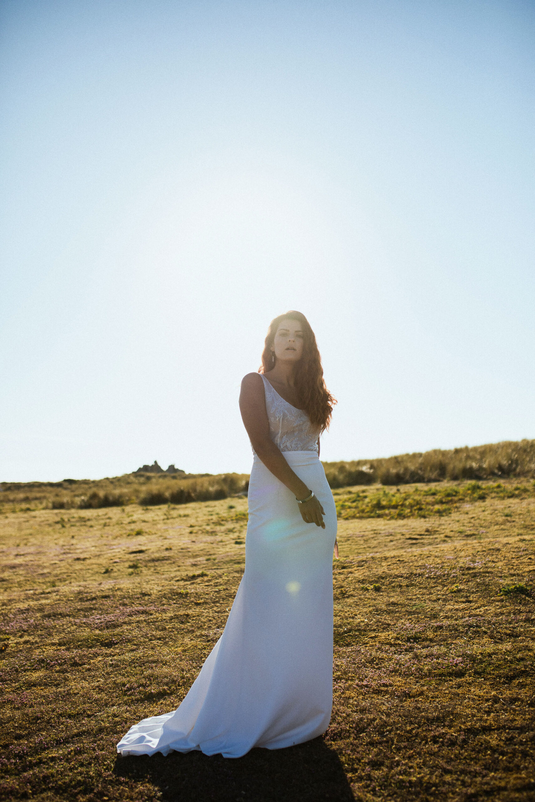 Femme rousse portant une robe de mariée en dentelle dans les dunes végétales en bord de mer.