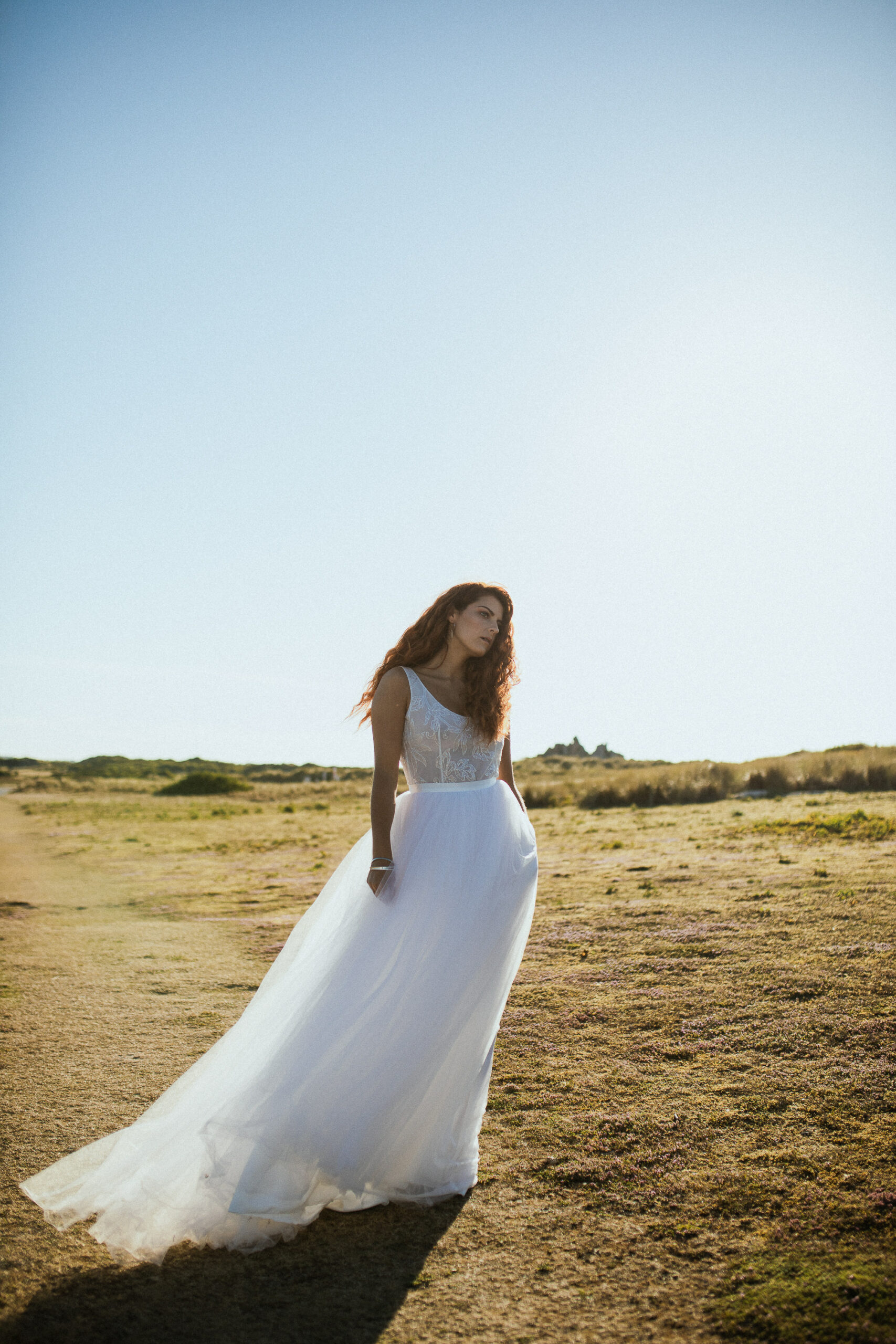 Femme rousse portant une robe de mariée en dentelle dans les dunes végétales en bord de mer.