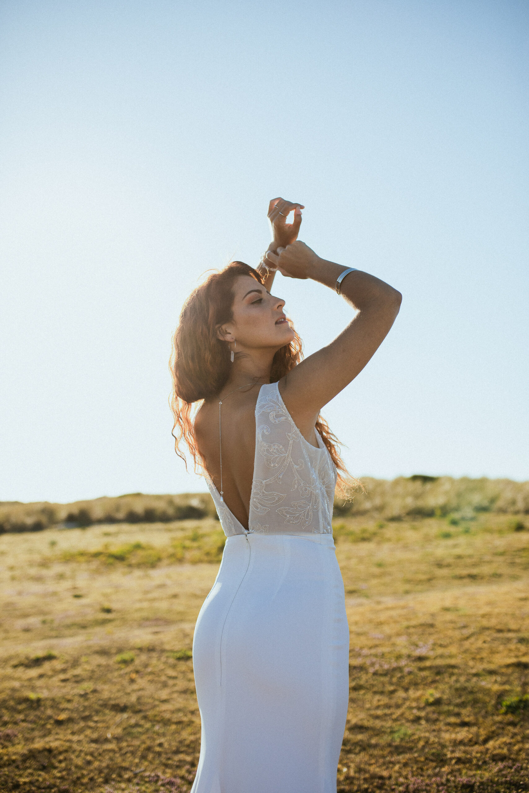 Femme rousse portant une robe de mariée en dentelle dans les dunes végétales en bord de mer.