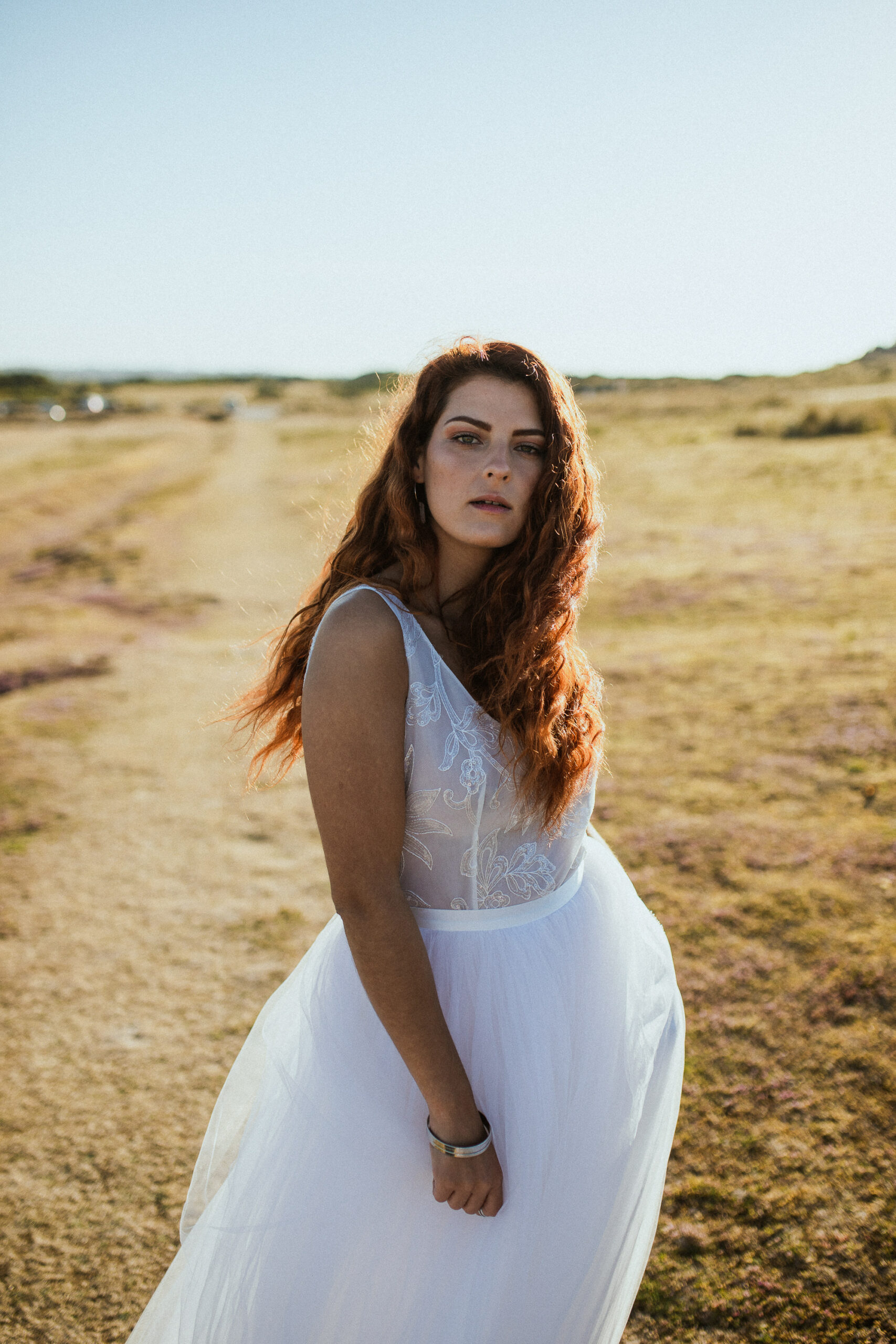 Femme rousse portant une robe de mariée en dentelle dans les dunes végétales en bord de mer.