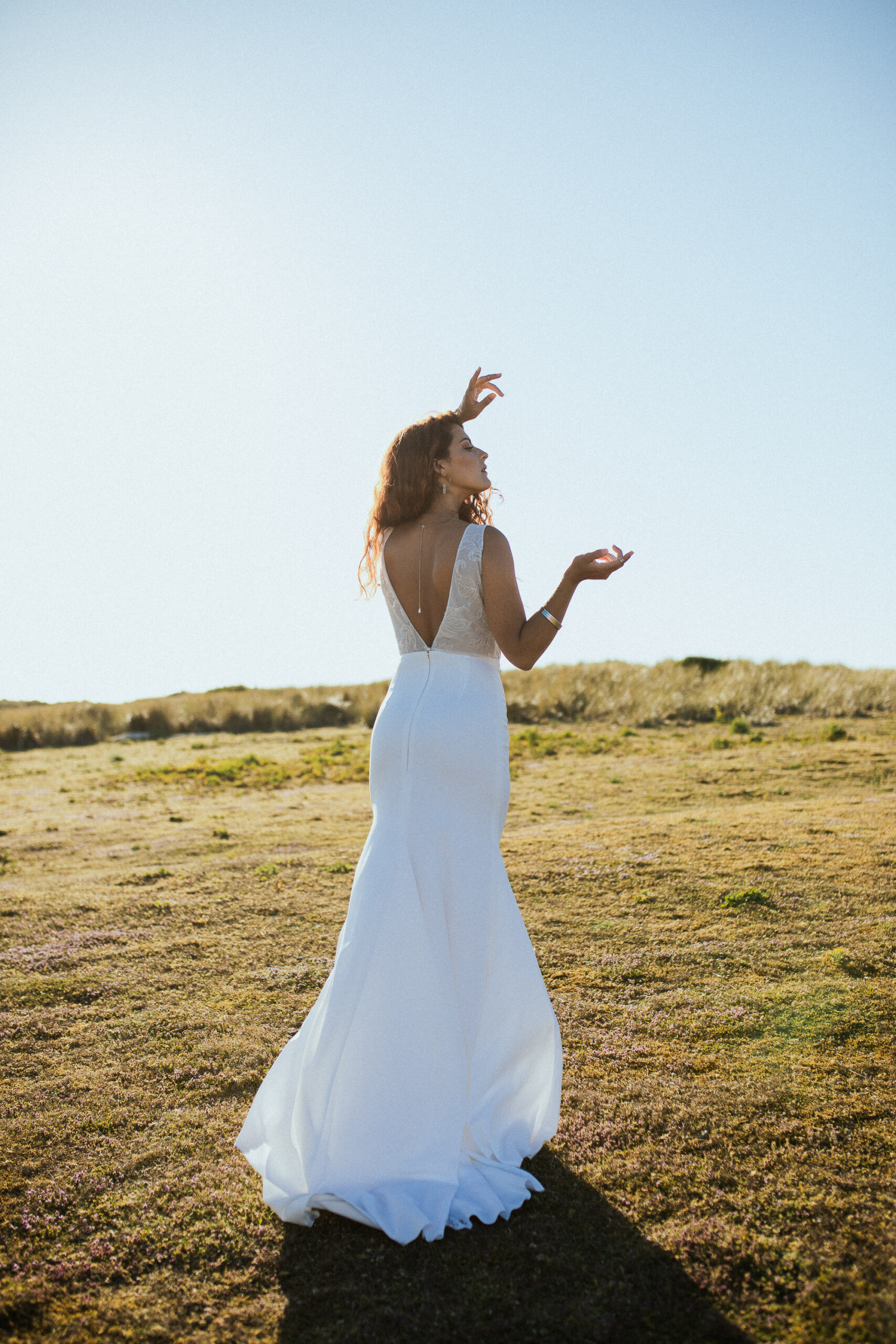 Femme rousse portant une robe de mariée en dentelle dans les dunes végétales en bord de mer.