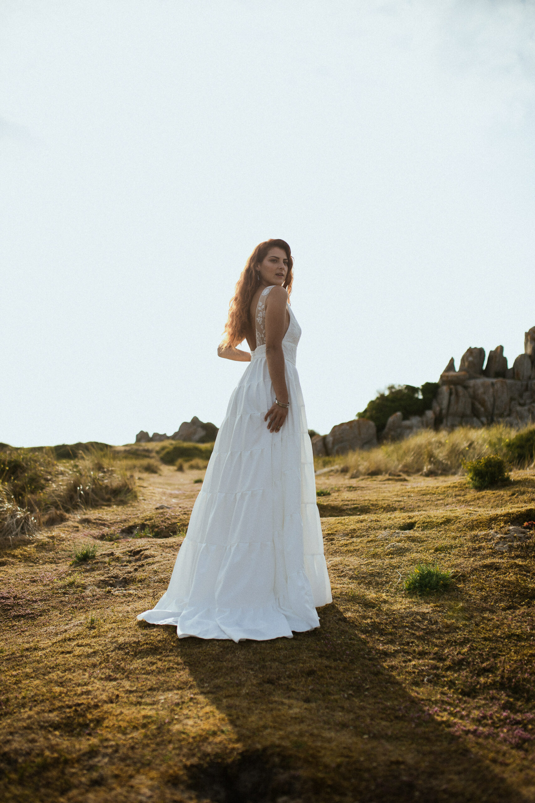 Femme rousse portant une robe de mariée en dentelle dans les dunes végétales en bord de mer.