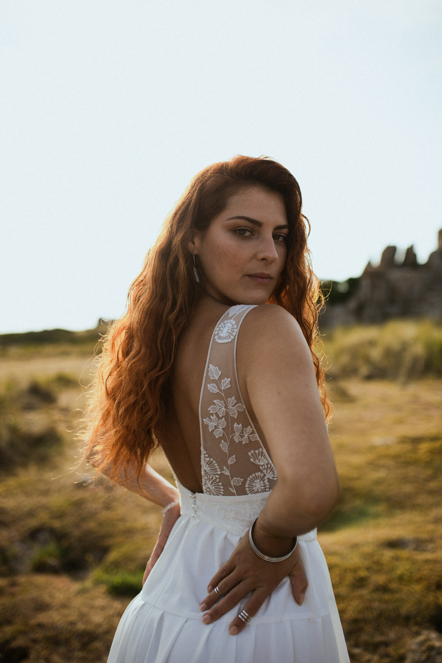 Femme rousse portant une robe de mariée en dentelle dans les dunes végétales en bord de mer.