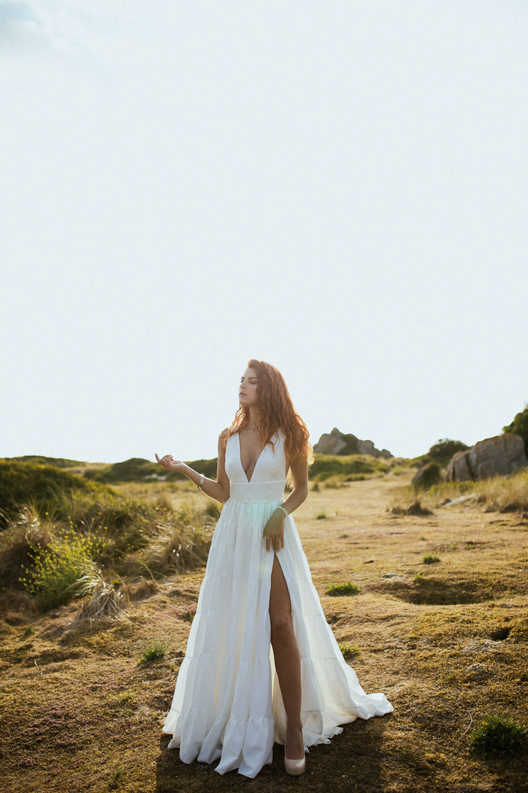 Femme rousse portant une robe de mariée en dentelle dans les dunes végétales en bord de mer.