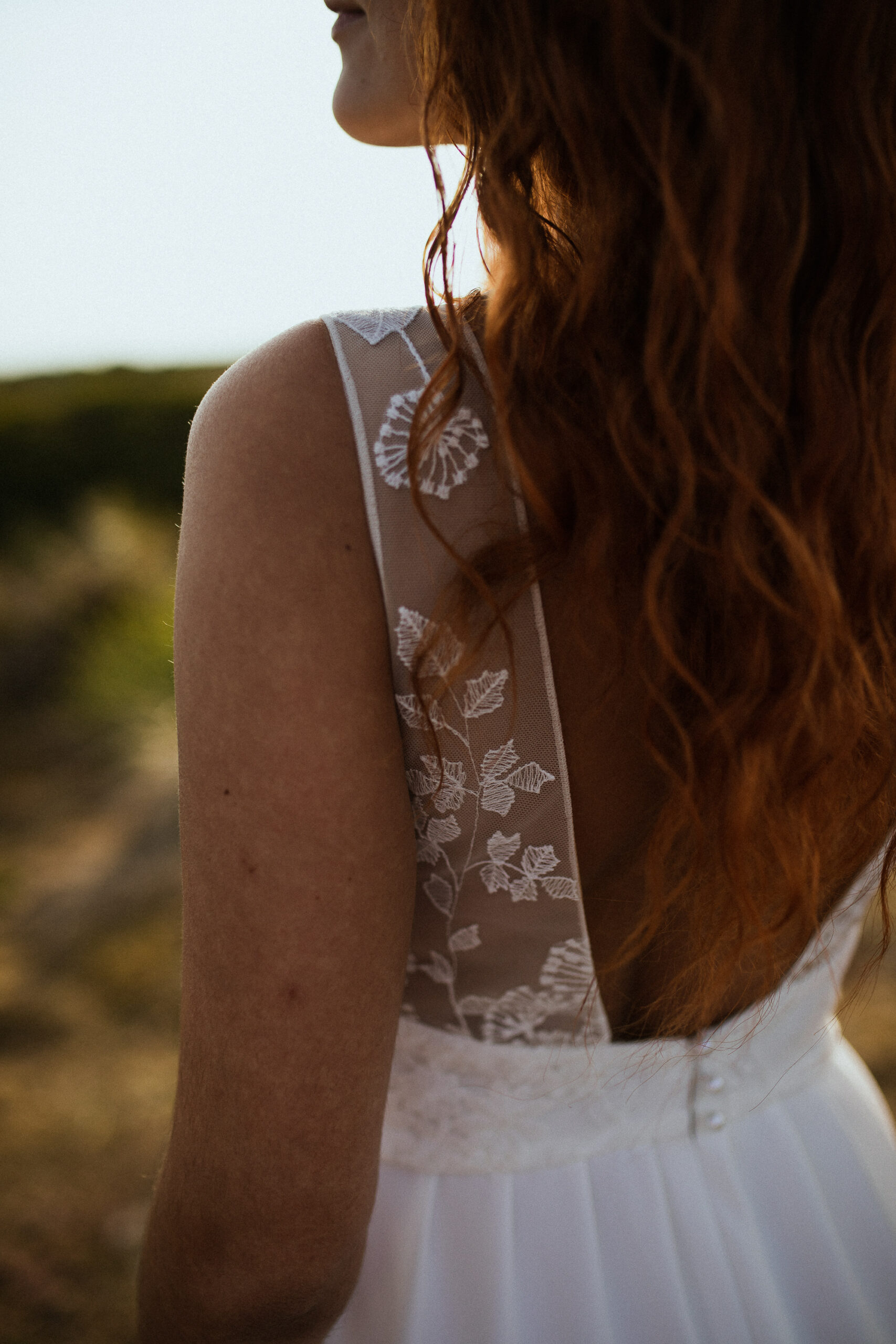 Femme rousse portant une robe de mariée en dentelle dans les dunes végétales en bord de mer.