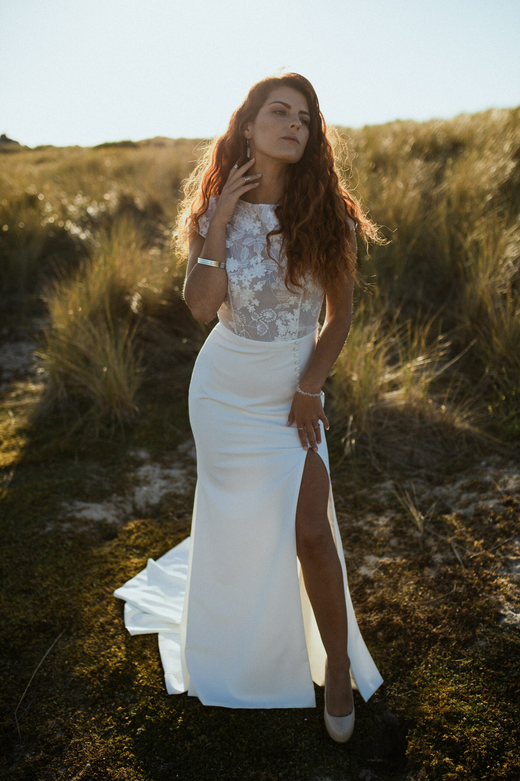 Femme rousse portant une robe de mariée en dentelle dans les dunes végétales en bord de mer.