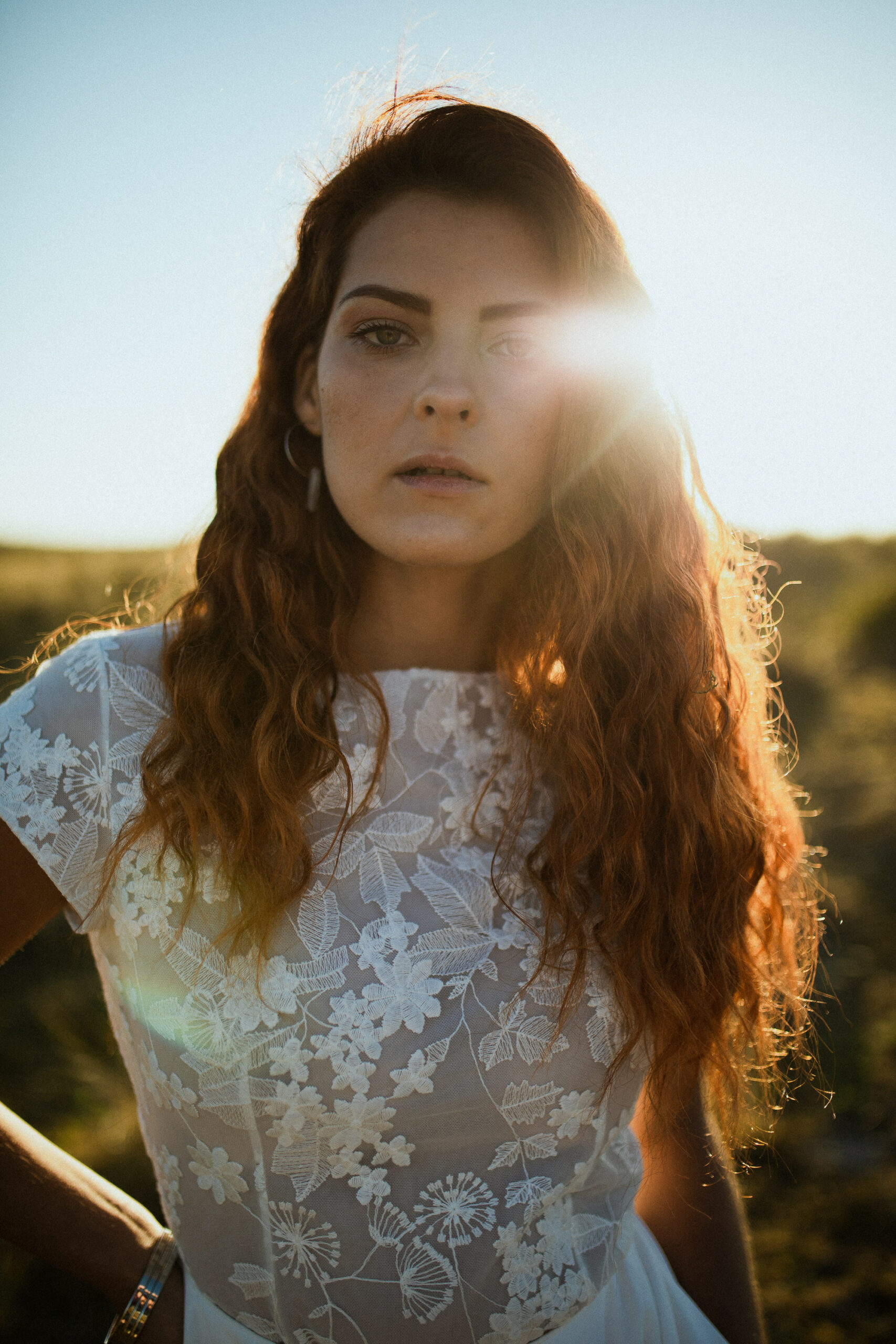 Femme rousse portant une robe de mariée en dentelle dans les dunes végétales en bord de mer.