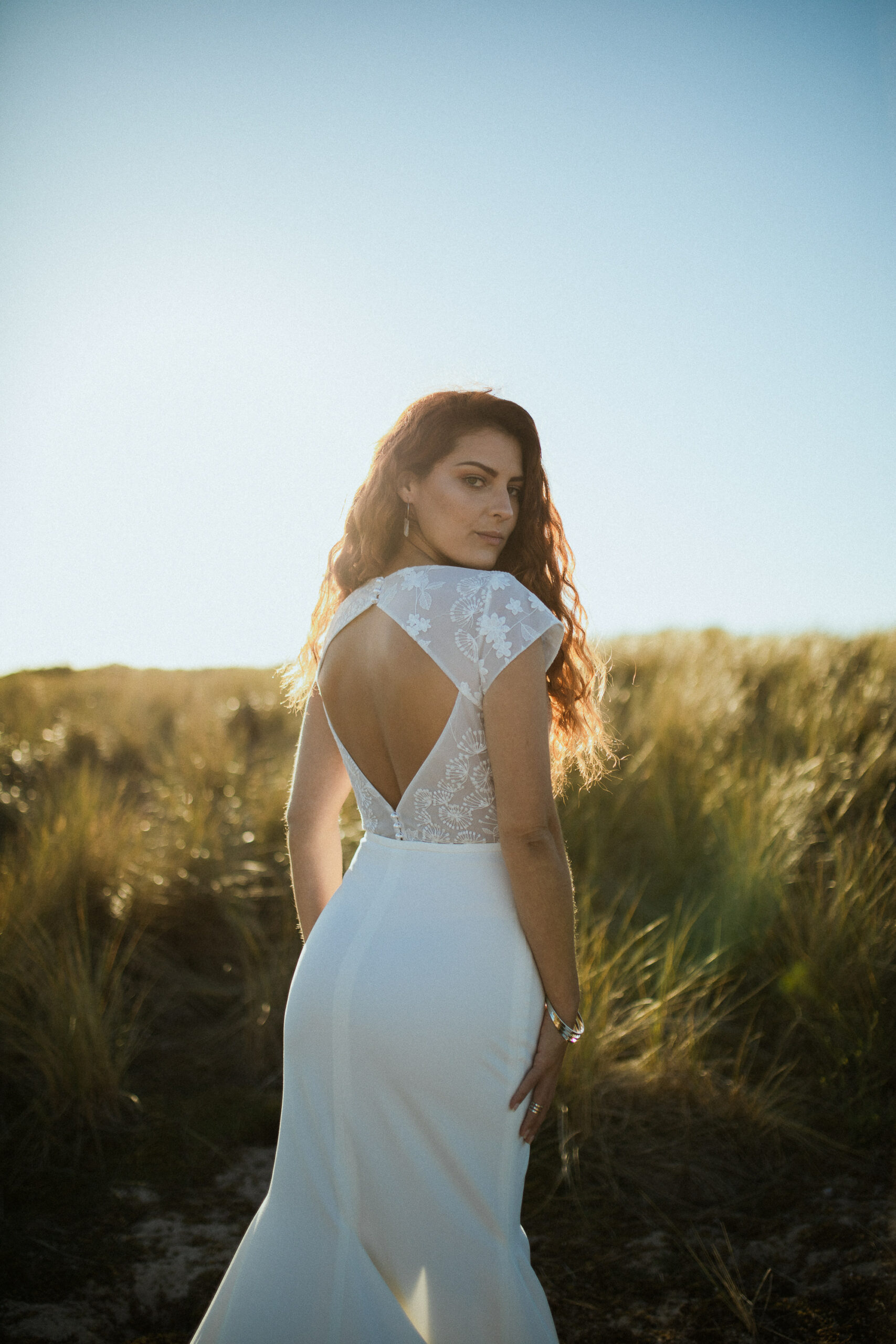 Femme rousse portant une robe de mariée en dentelle dans les dunes végétales en bord de mer.