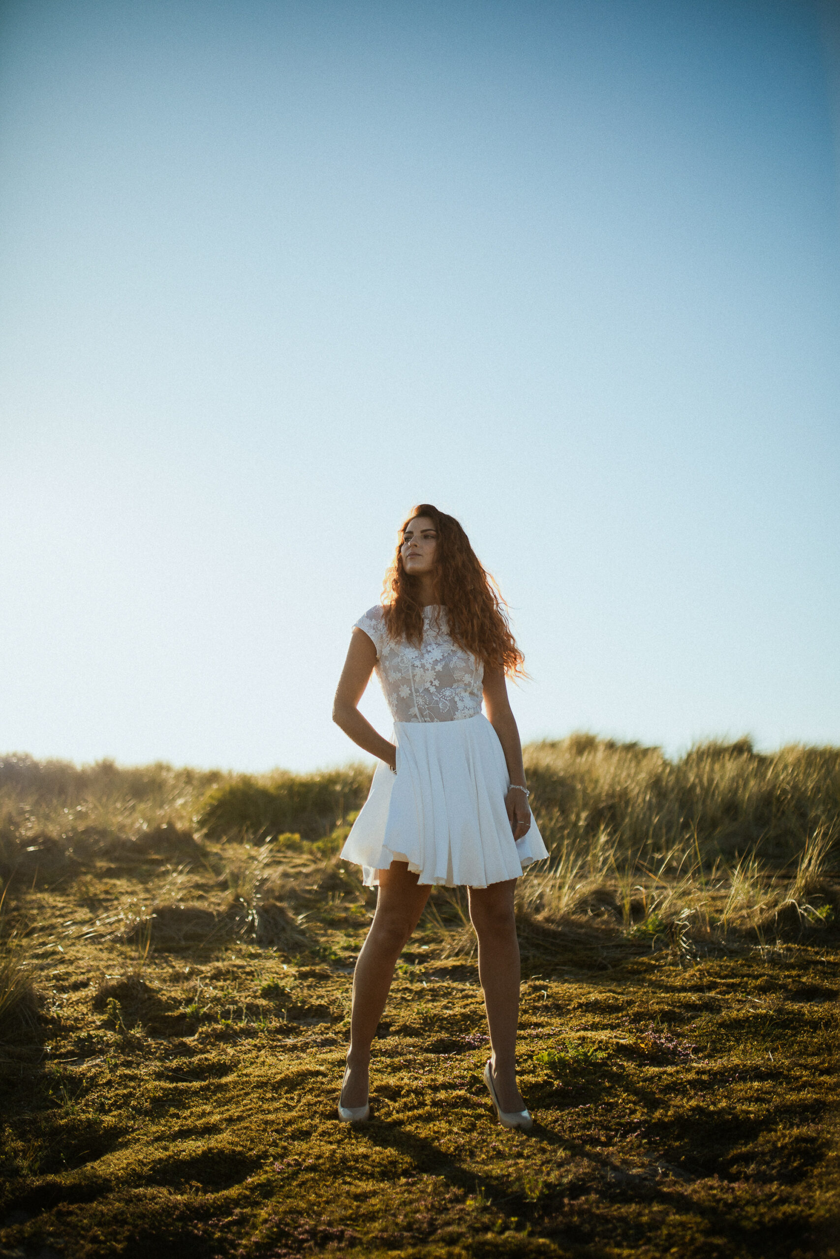 Femme rousse portant une robe de mariée en dentelle dans les dunes végétales en bord de mer.