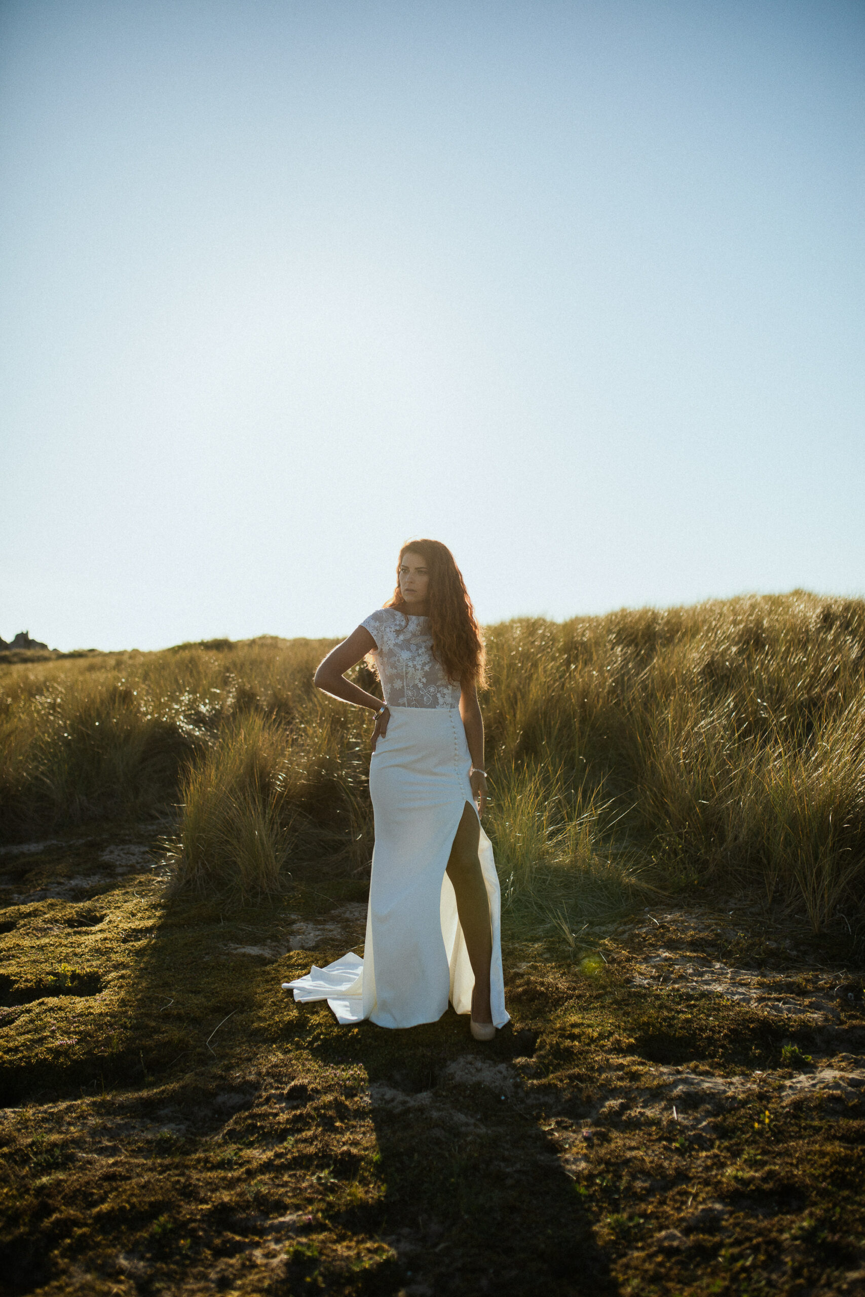 Femme rousse portant une robe de mariée en dentelle dans les dunes végétales en bord de mer.