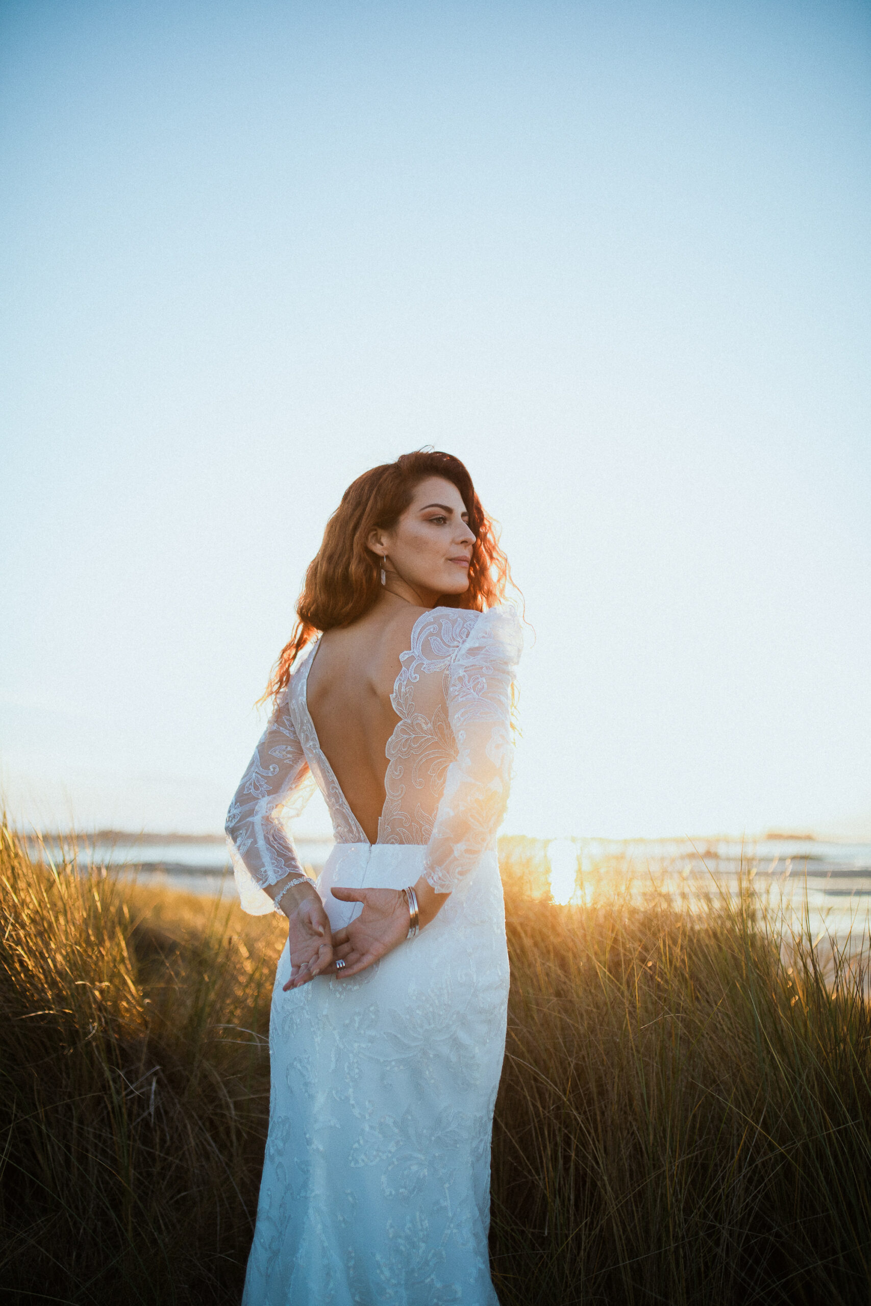 Femme rousse portant une robe de mariée en dentelle dans les dunes végétales en bord de mer.