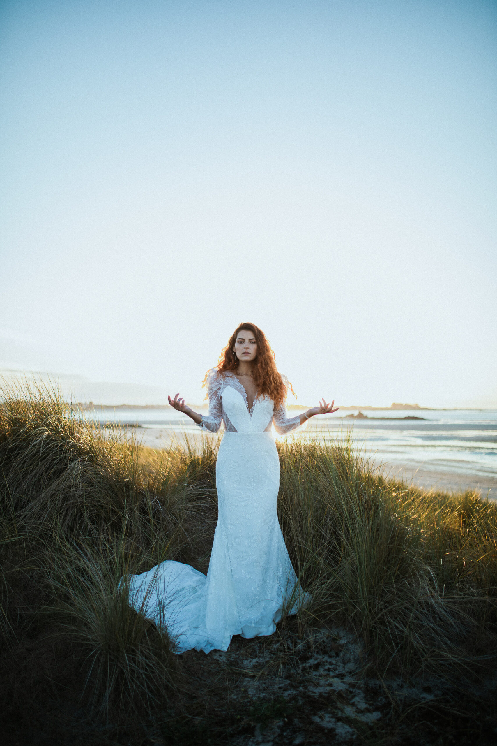 Femme rousse portant une robe de mariée en dentelle dans les dunes végétales en bord de mer.