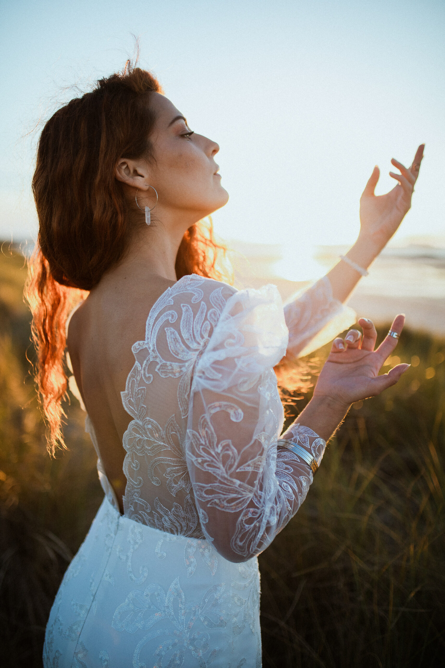 Femme rousse portant une robe de mariée en dentelle dans les dunes végétales en bord de mer.