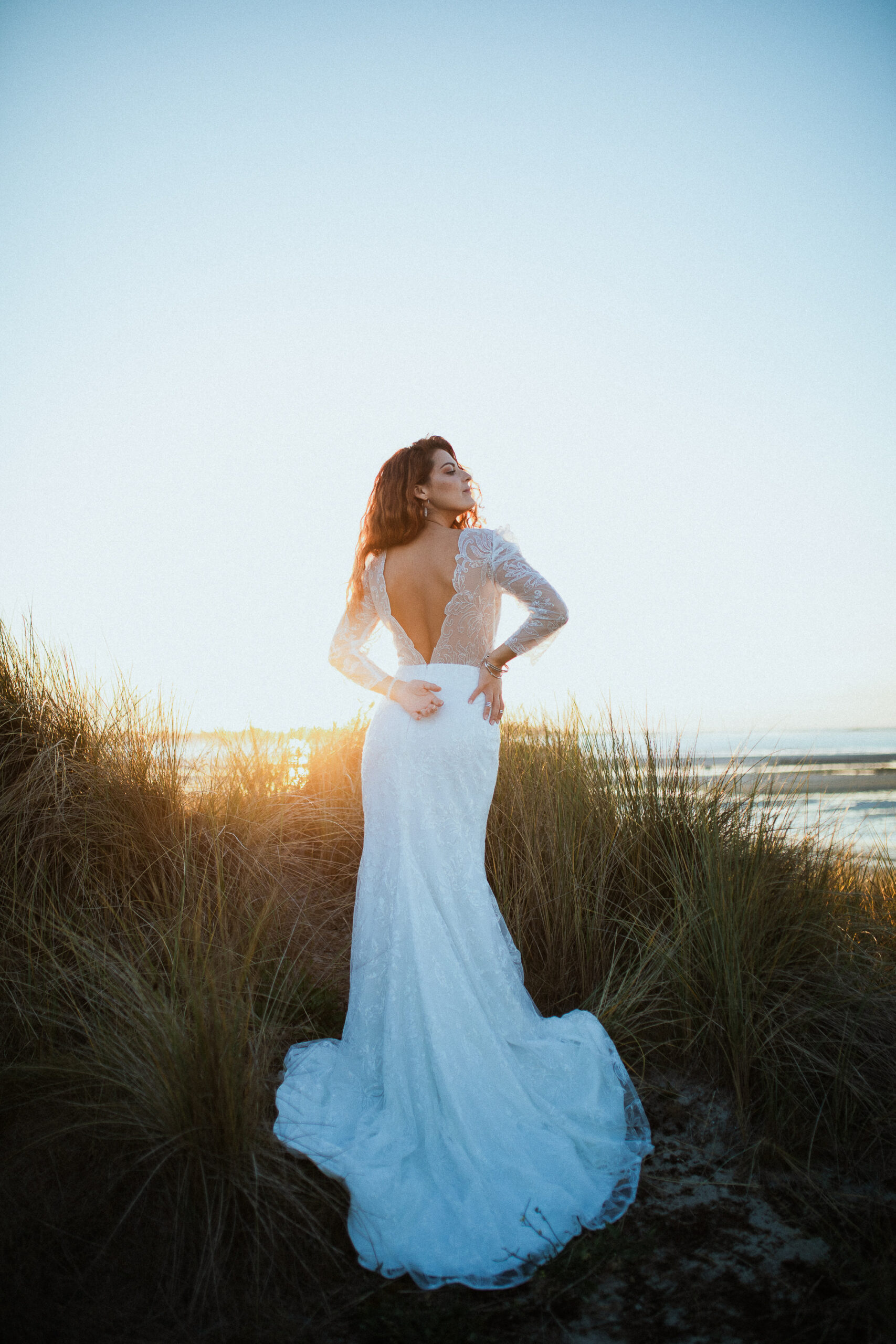 Femme rousse portant une robe de mariée en dentelle dans les dunes végétales en bord de mer.