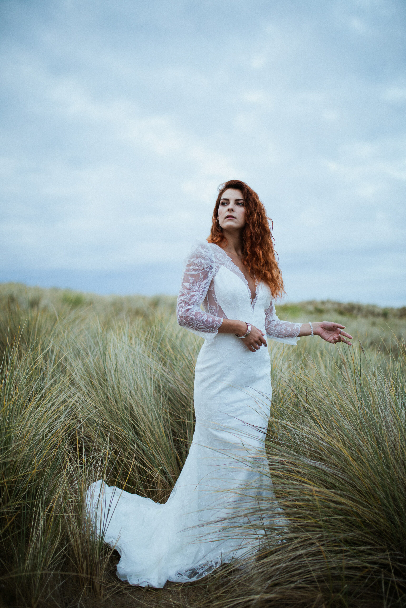 Femme rousse portant une robe de mariée en dentelle dans les dunes végétales en bord de mer.