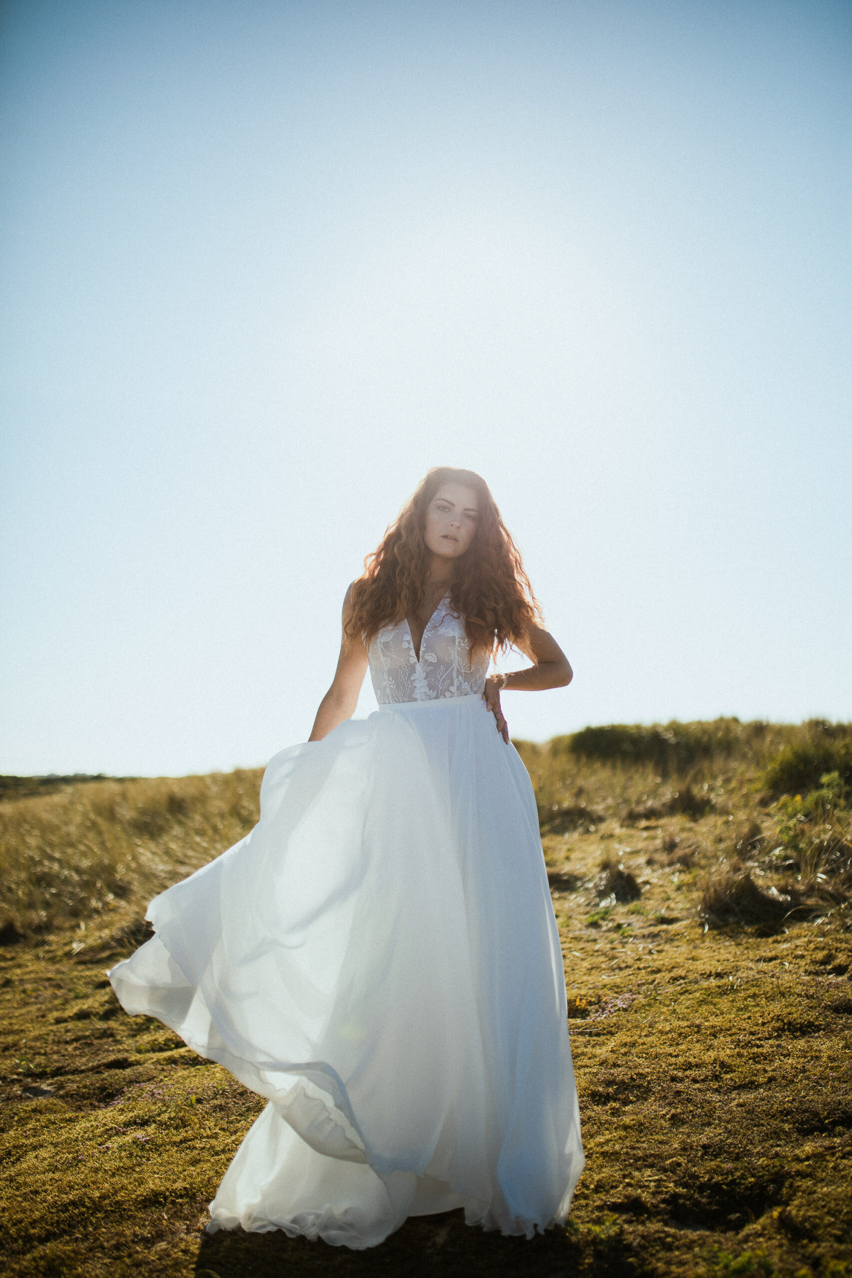 Femme rousse portant une robe de mariée en dentelle dans les dunes végétales en bord de mer.