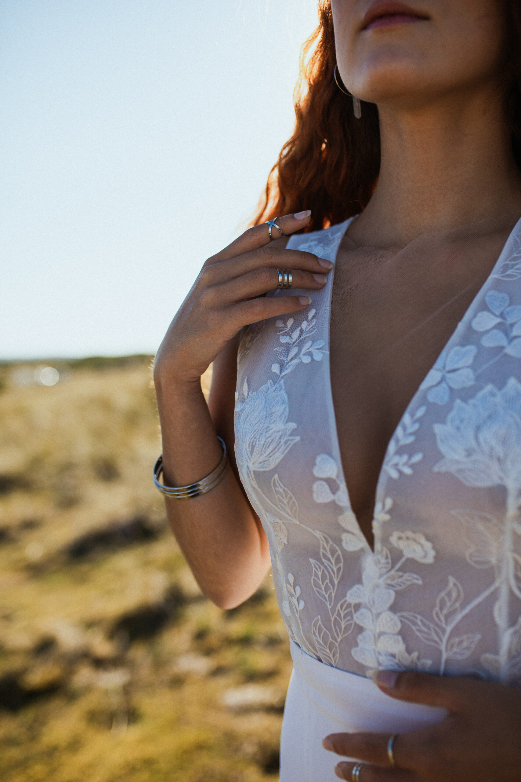 Femme rousse portant une robe de mariée en dentelle dans les dunes végétales en bord de mer.