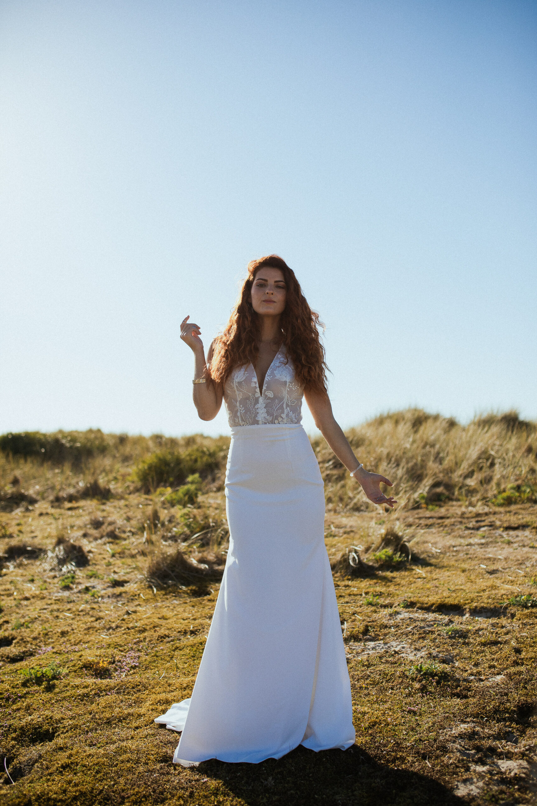 Femme rousse portant une robe de mariée en dentelle dans les dunes végétales en bord de mer.