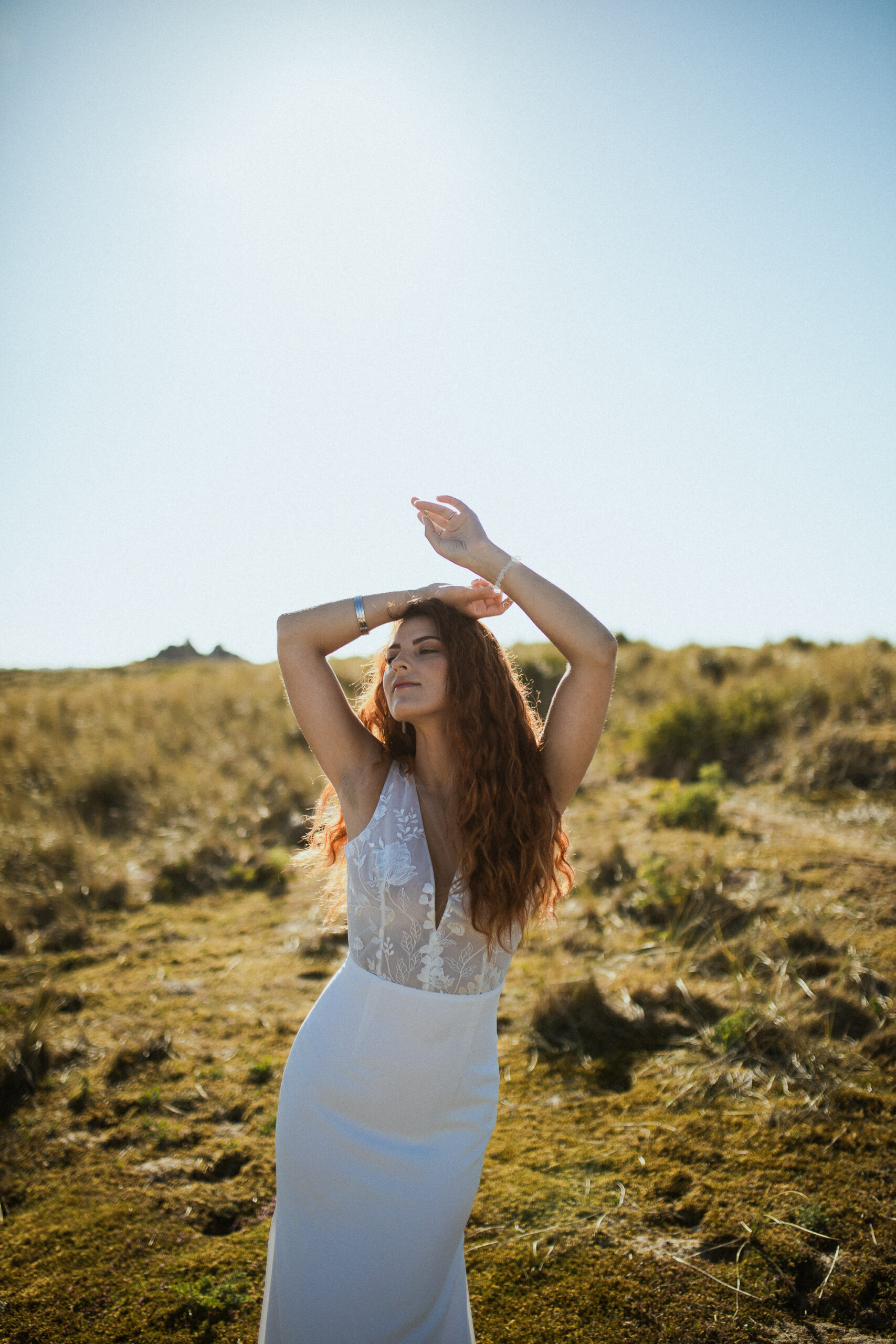 Femme rousse portant une robe de mariée en dentelle dans les dunes végétales en bord de mer.