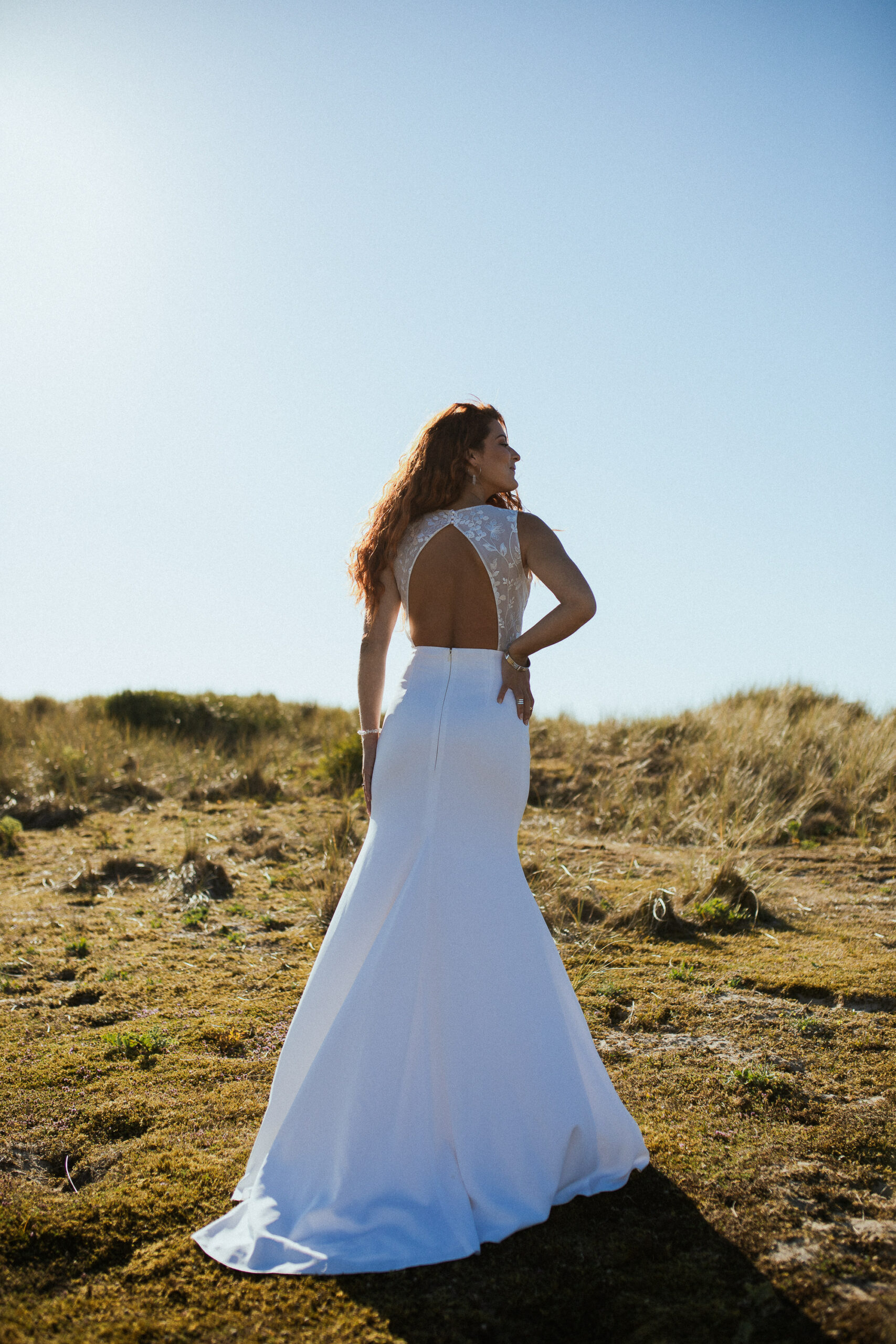 Femme rousse portant une robe de mariée en dentelle dans les dunes végétales en bord de mer.