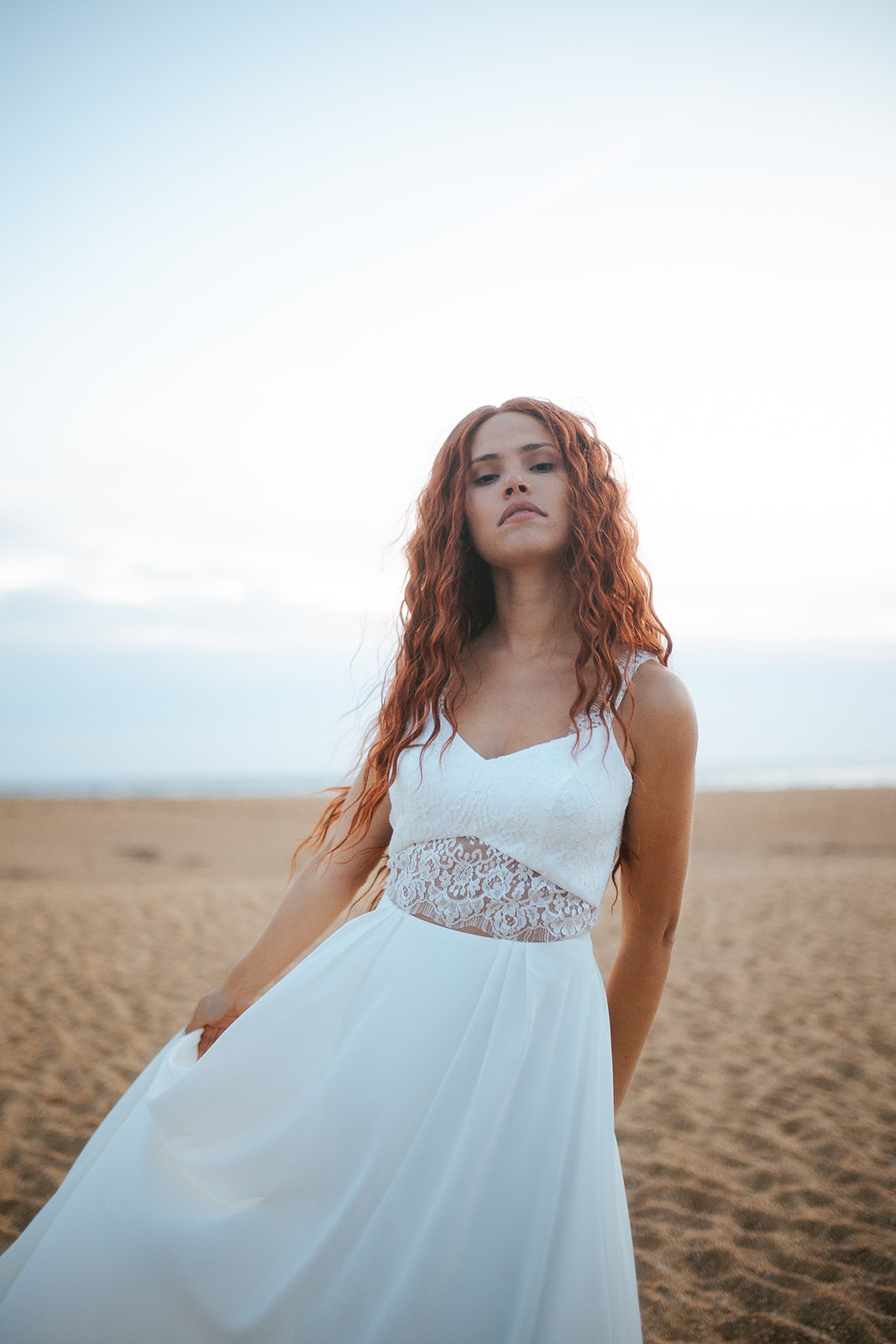 Femme rousse portant une robe de mariée de créateur en dentelle sur-mesure à la plage.