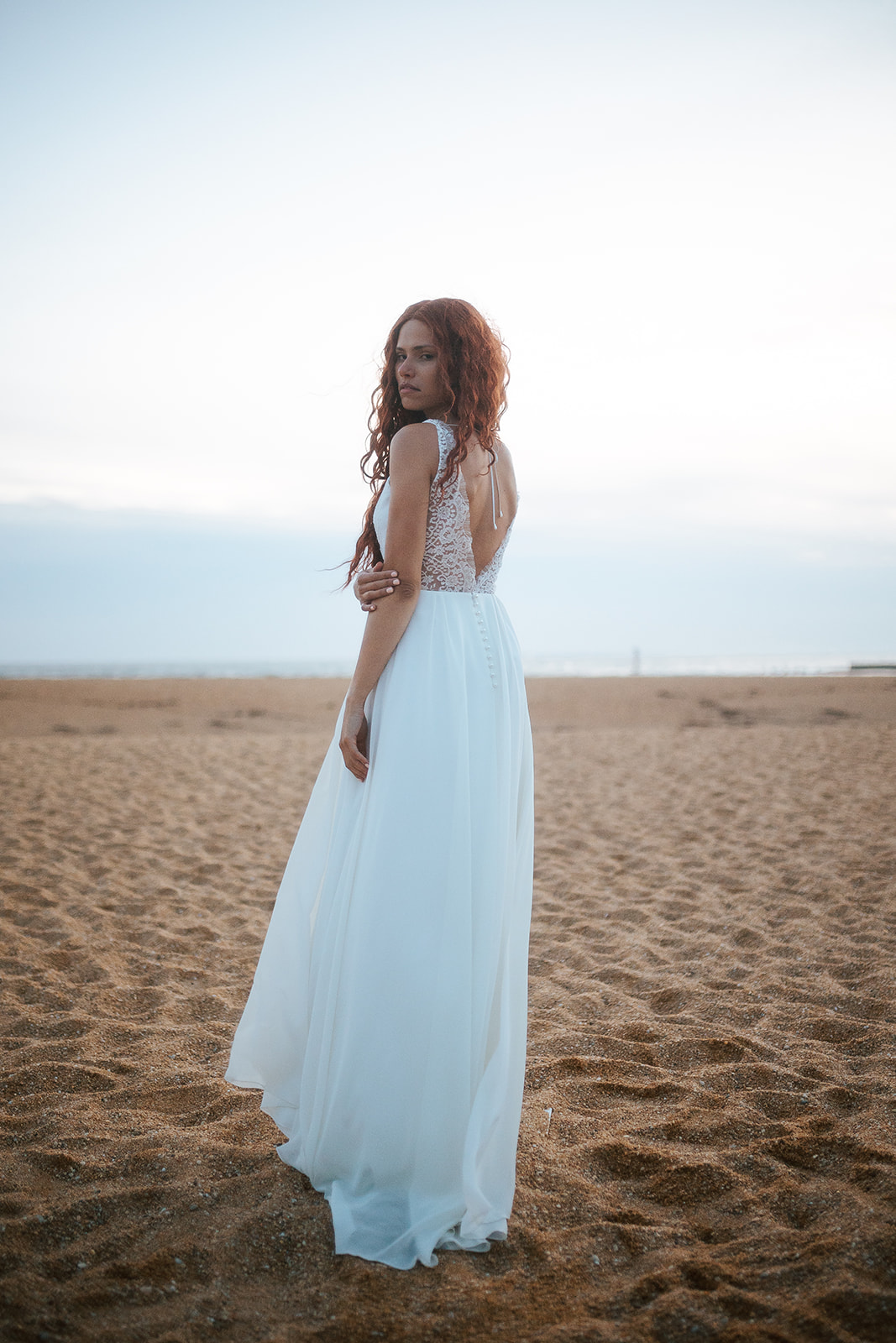 Femme rousse portant une robe de mariée de créateur en dentelle sur-mesure à la plage.