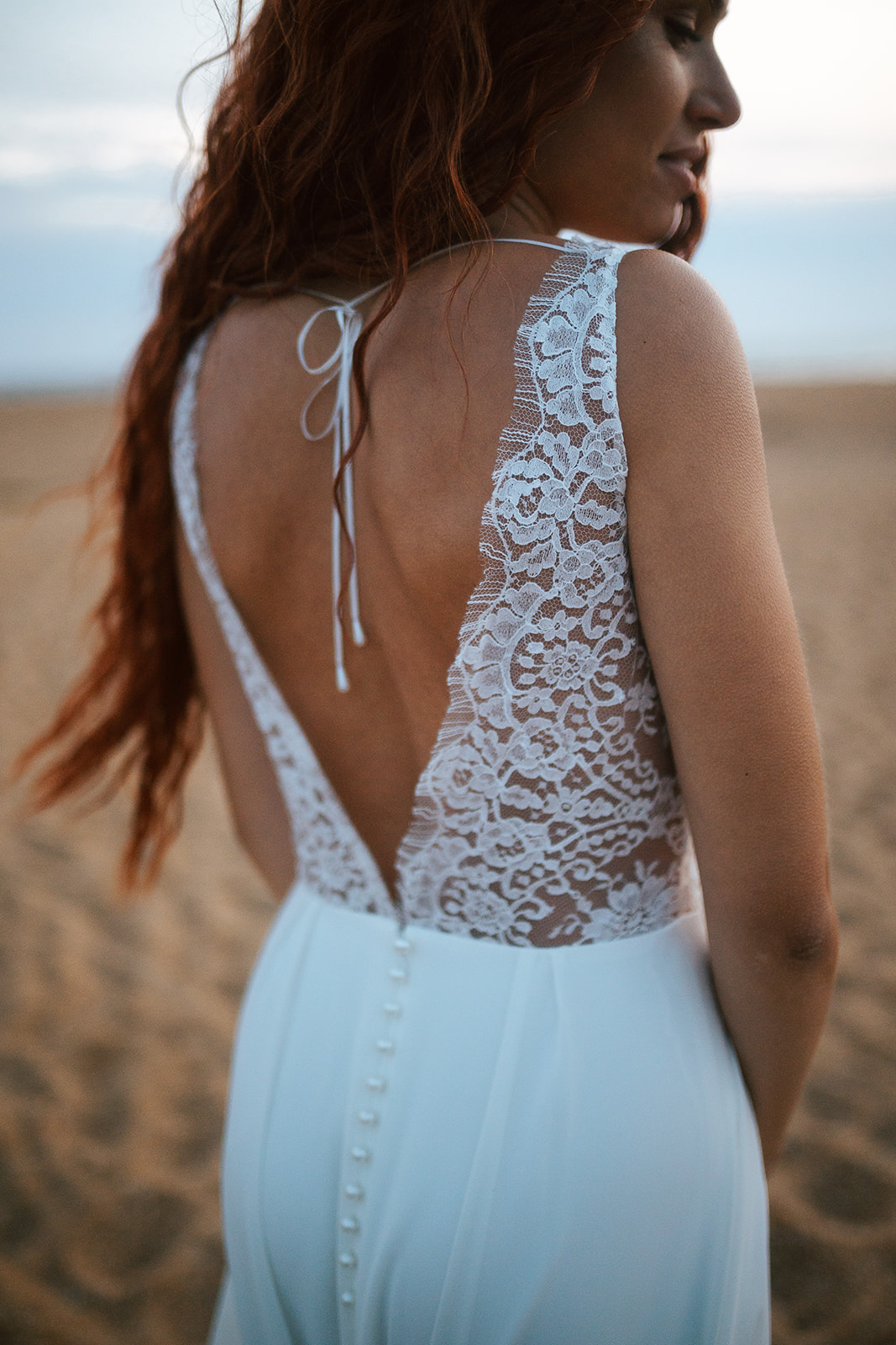 Femme rousse portant une robe de mariée de créateur en dentelle sur-mesure à la plage.
