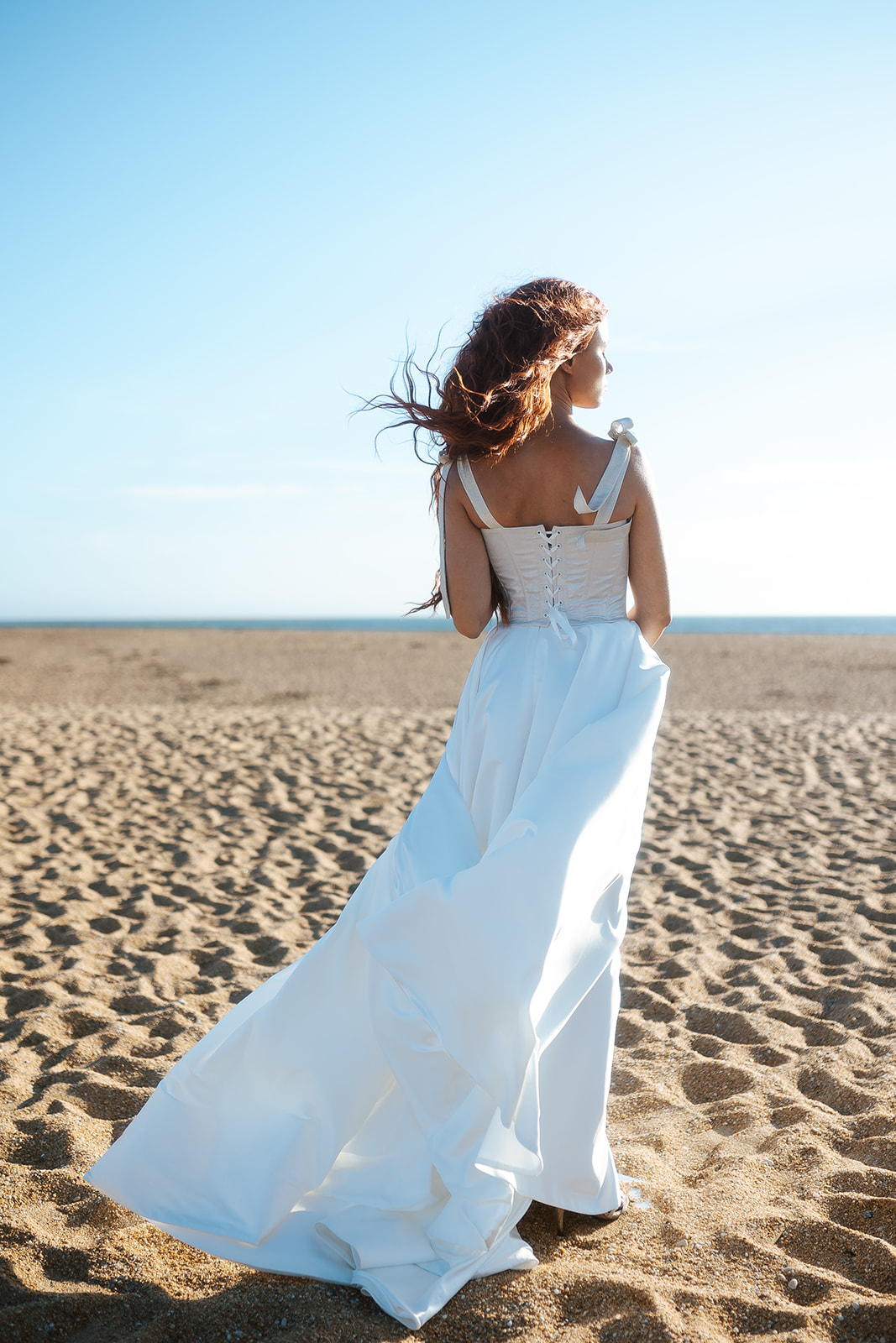 Femme rousse portant une robe de mariée de créateur en dentelle sur-mesure à la plage.