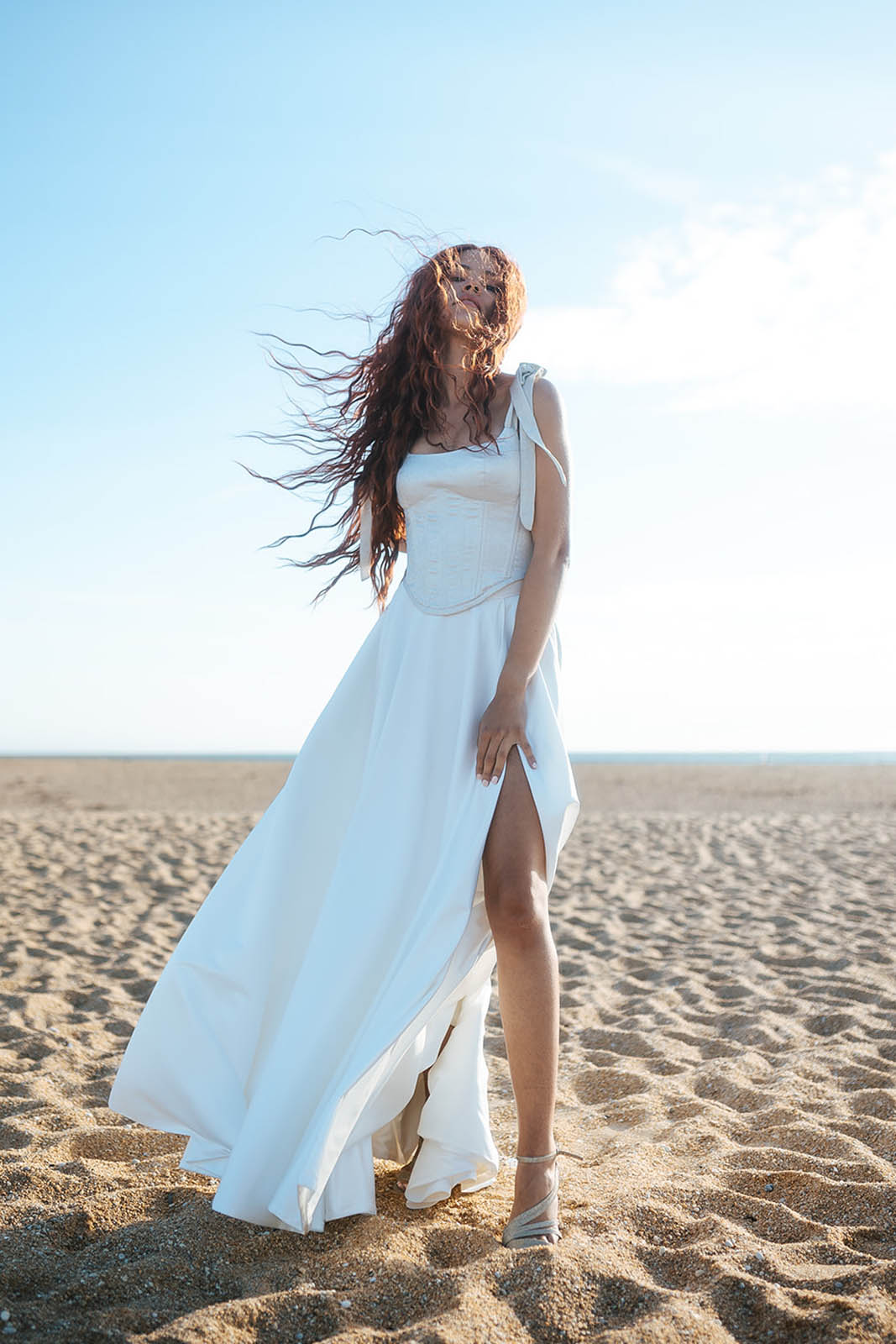 Femme rousse portant une robe de mariée de créateur en dentelle sur-mesure à la plage.