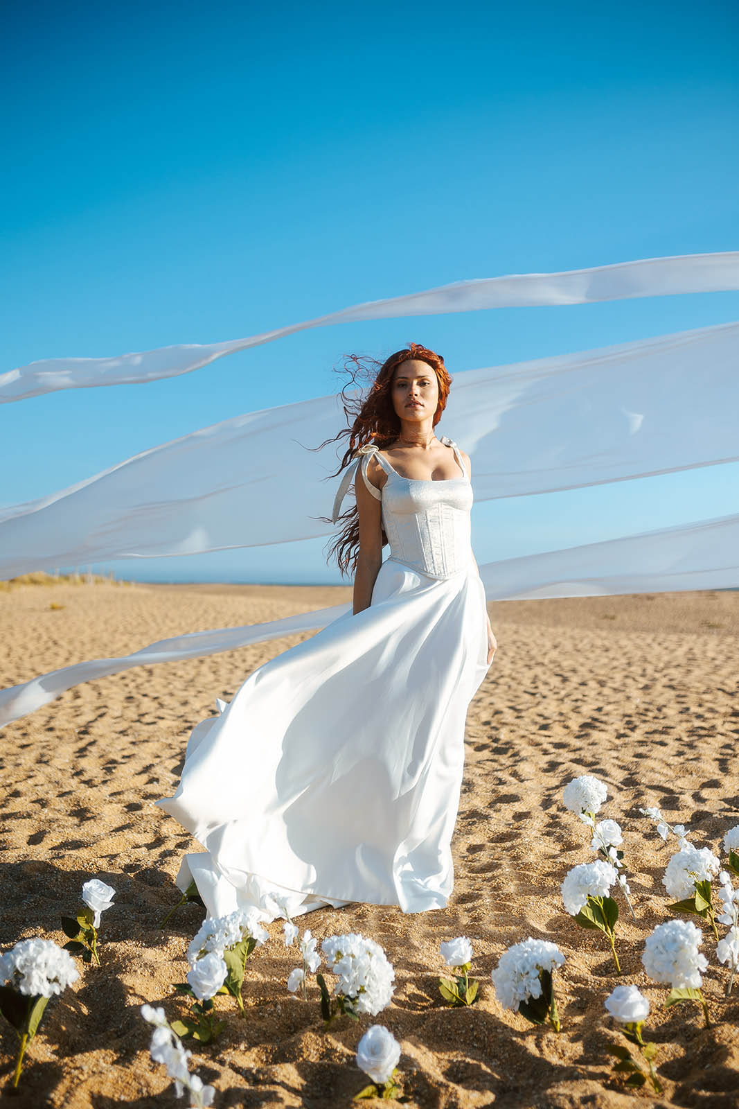 Femme rousse portant une robe de mariée de créateur en dentelle sur-mesure à la plage.