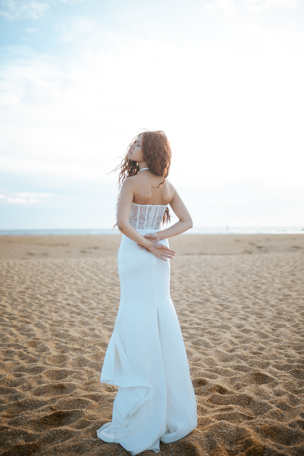Femme rousse portant une robe de mariée de créateur en dentelle sur-mesure à la plage.