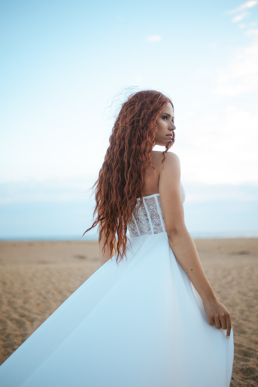 Femme rousse portant une robe de mariée de créateur en dentelle sur-mesure à la plage.