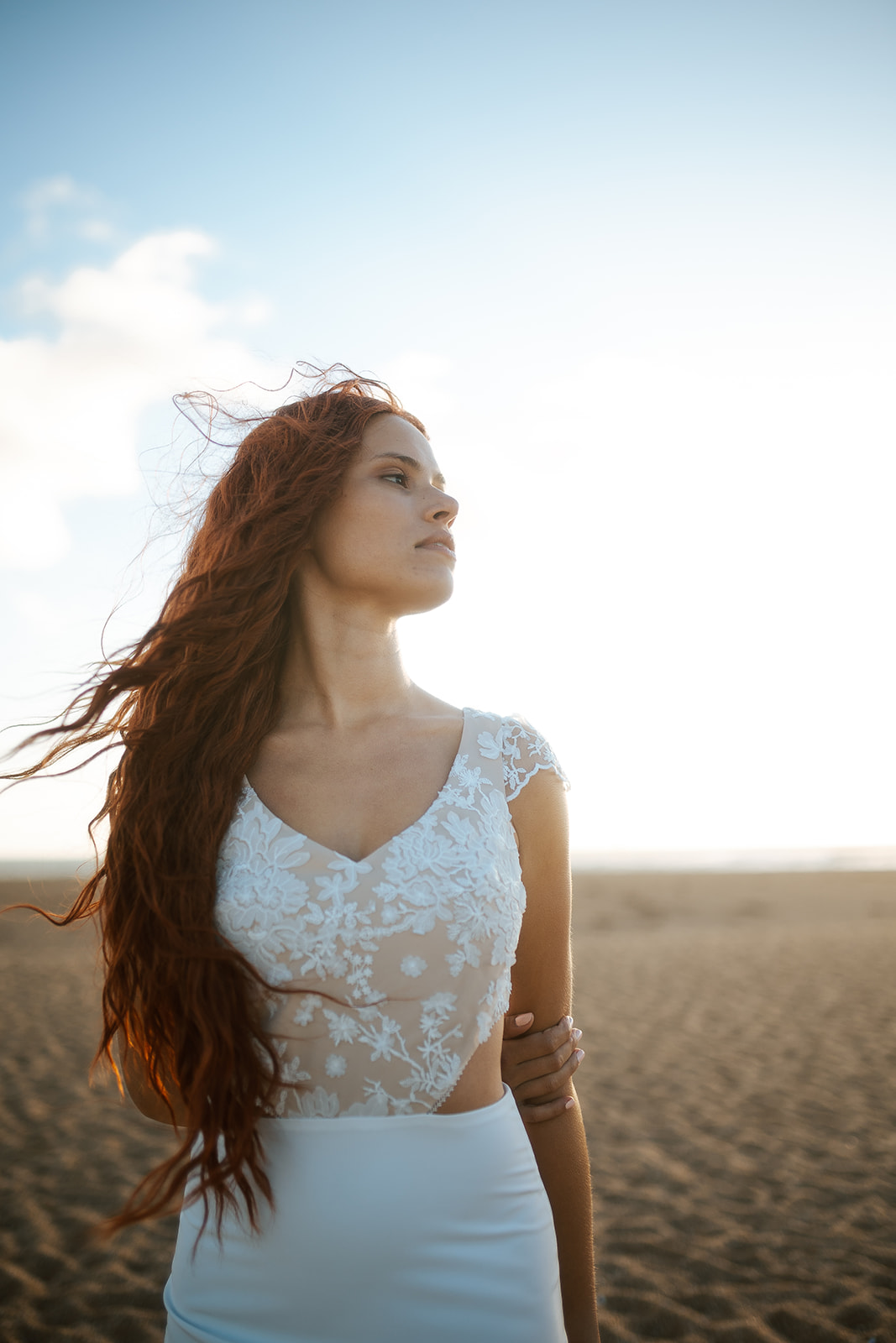 Femme rousse portant une robe de mariée de créateur en dentelle sur-mesure à la plage.