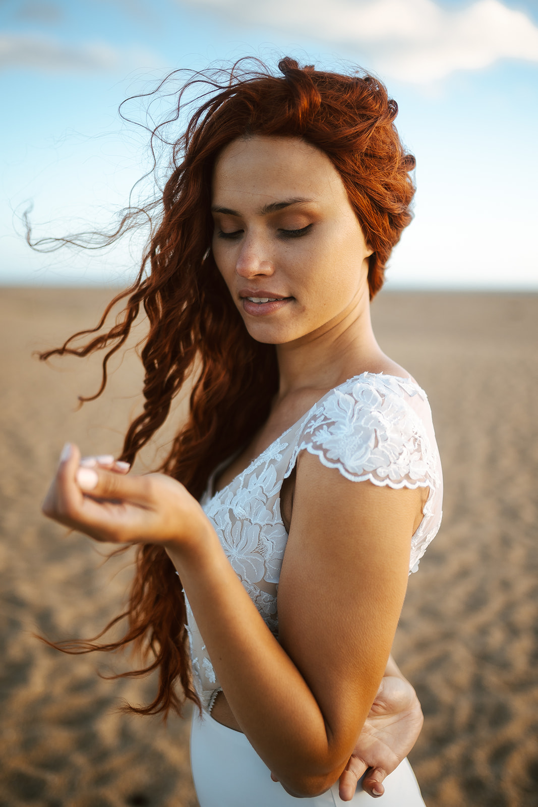 Femme rousse portant une robe de mariée de créateur en dentelle sur-mesure à la plage.