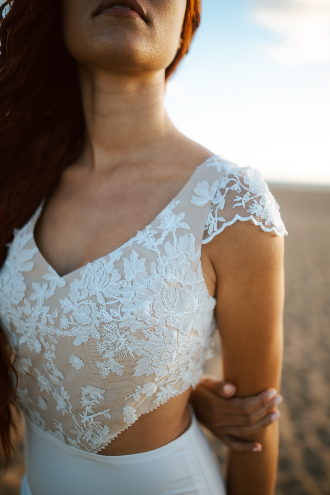 Femme rousse portant une robe de mariée de créateur en dentelle sur-mesure à la plage.