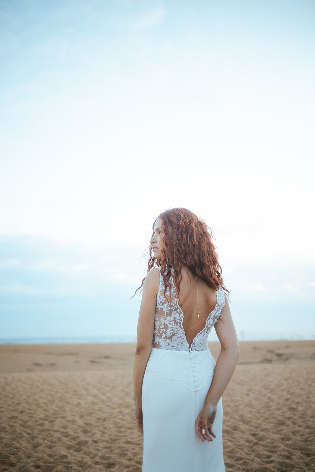 Femme rousse portant une robe de mariée de créateur en dentelle sur-mesure à la plage.