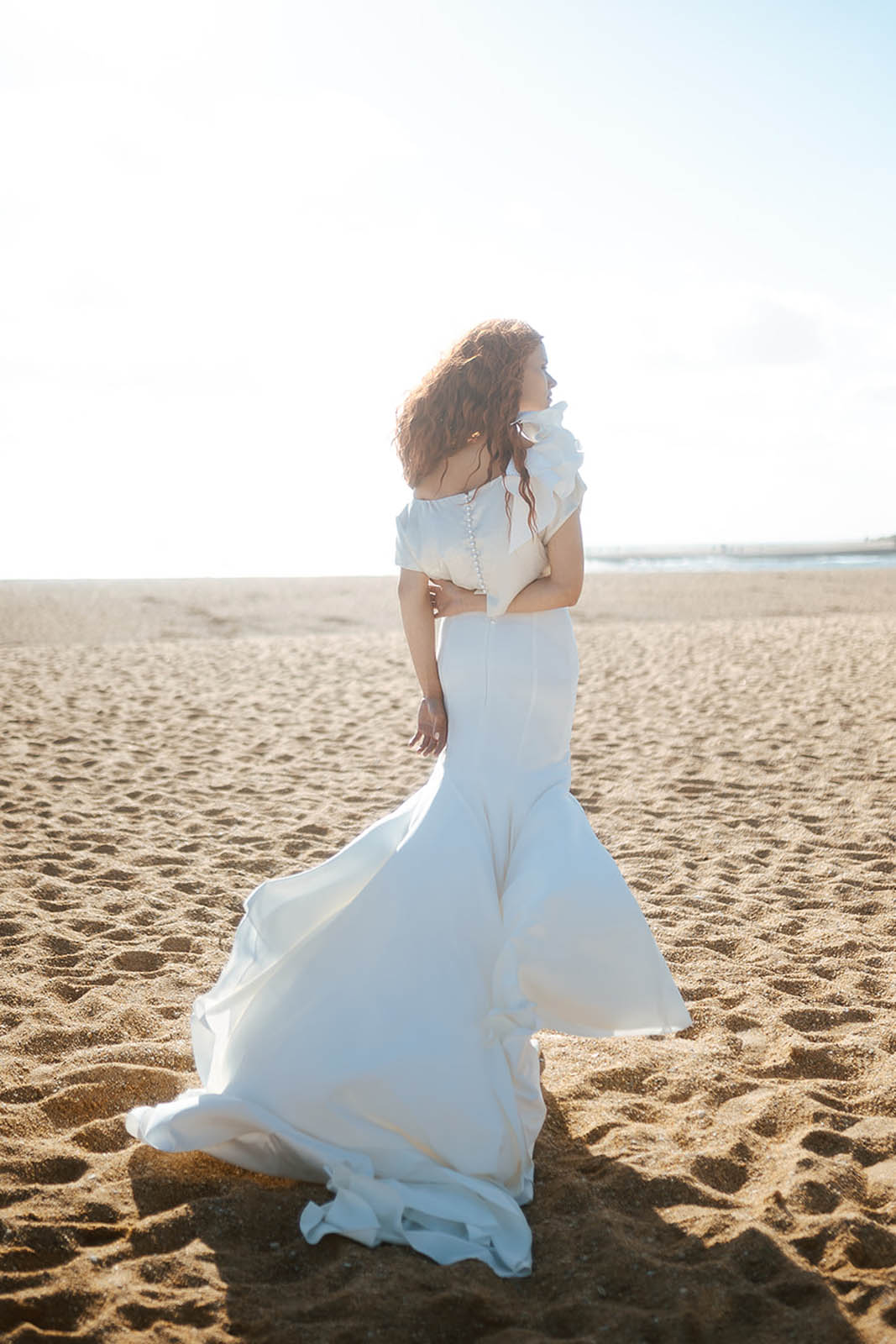 Femme rousse portant une robe de mariée de créateur en dentelle sur-mesure à la plage.
